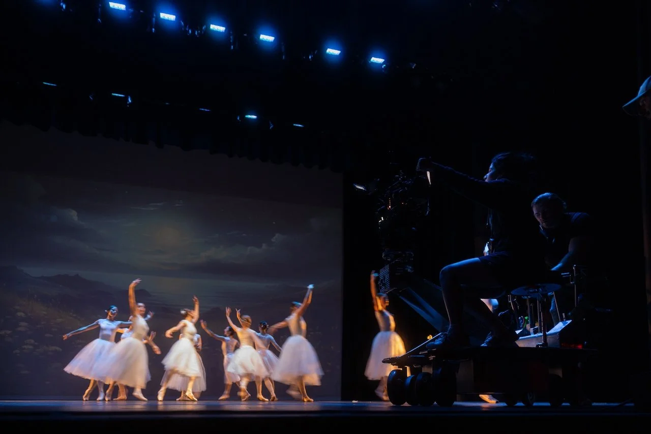 Ballet dancers performing on stage with a scenic backdrop, camera operators filming the performance, stage lights overhead.