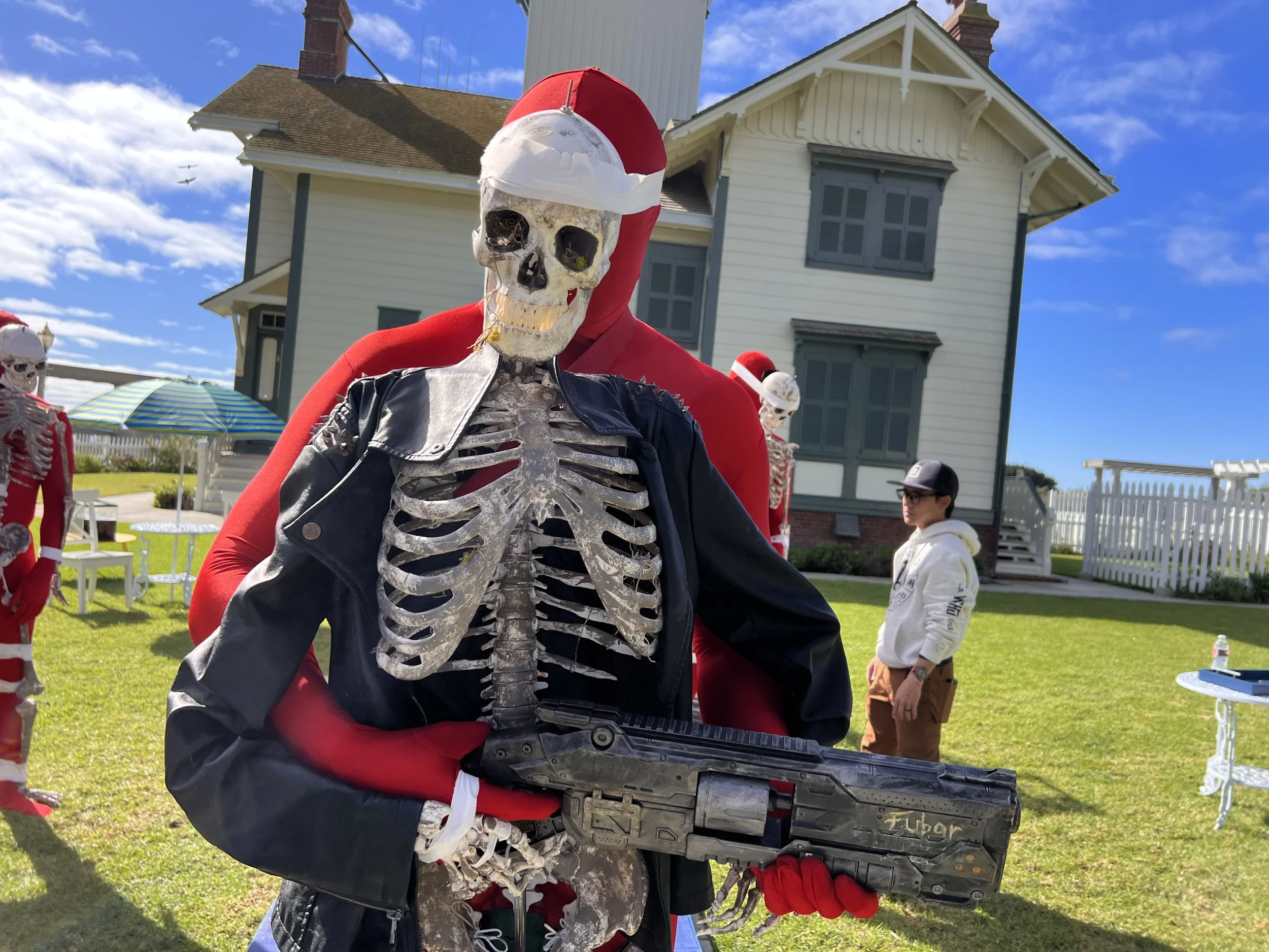 Skeleton dressed in black jacket with skeleton ribs, wearing a Santa hat and red gloves, holding a toy gun, outdoors in front of a house with other skeleton decorations and a person wearing a hoodie and cap.