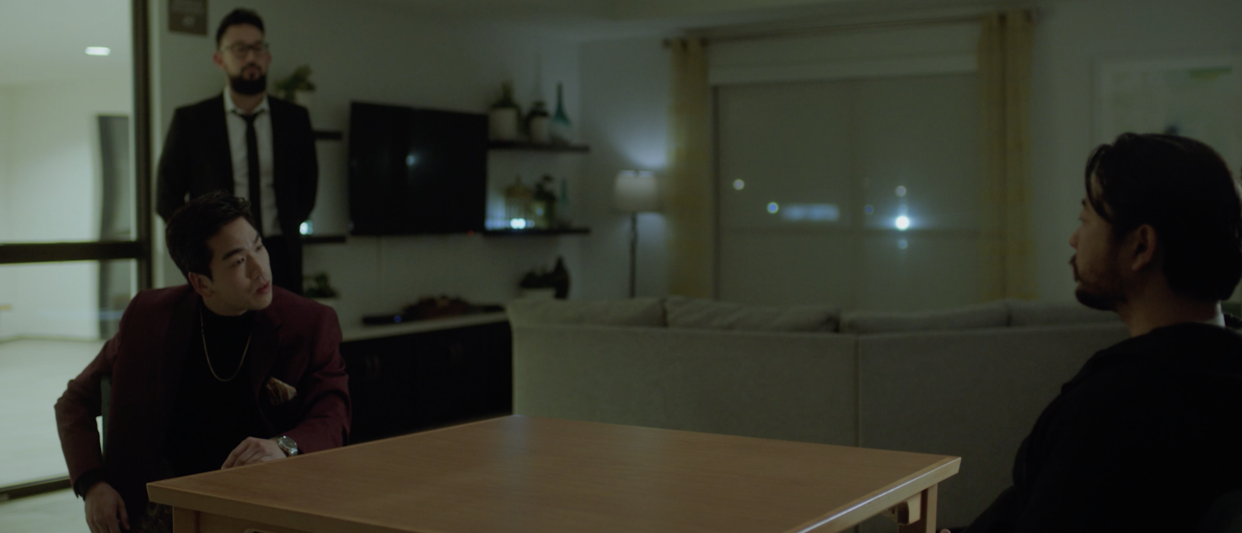 Three men in a dimly lit living room having a serious conversation, with one man standing in the background and two men sitting at a table in the foreground.