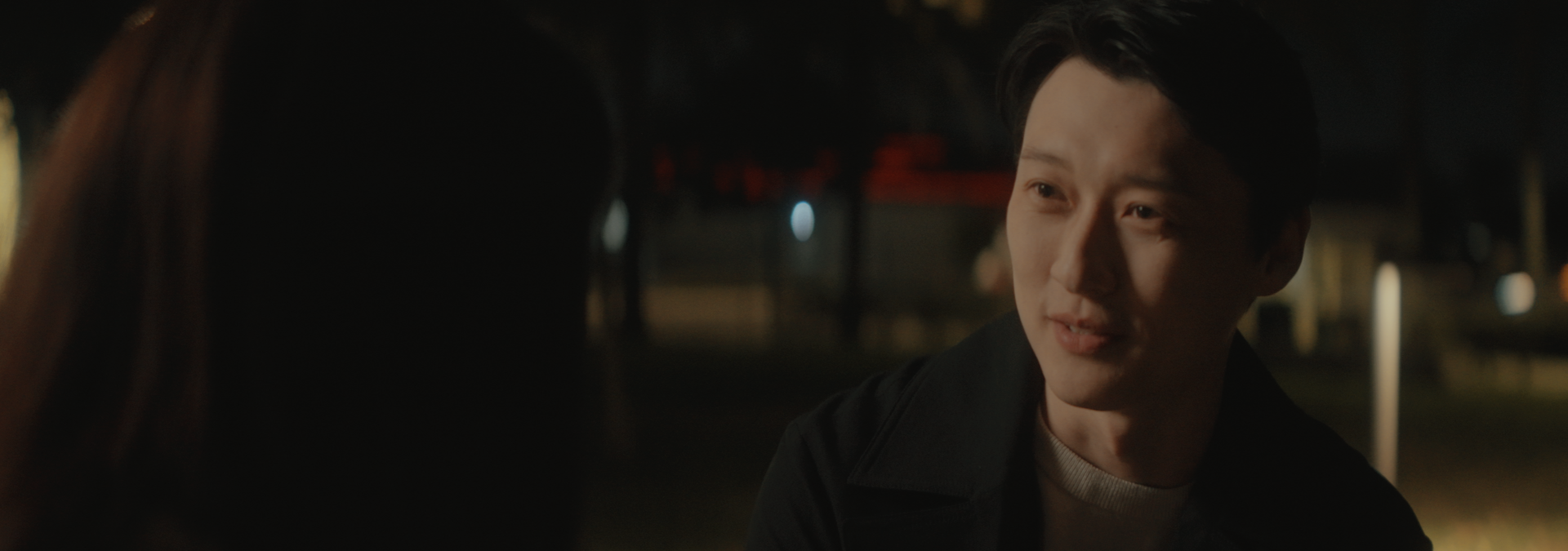 A young man and woman having a conversation outdoors at night, with a dimly lit background.