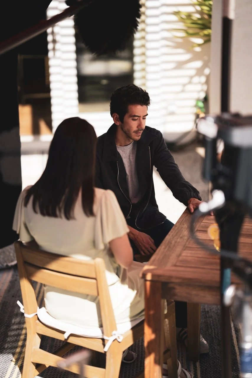 A man and woman sit at a wooden table in a well-lit room, engaged in a conversation. The man has dark, curly hair and is wearing a black jacket and gray shirt, while the woman has straight, dark hair and is wearing a light-colored blouse.
