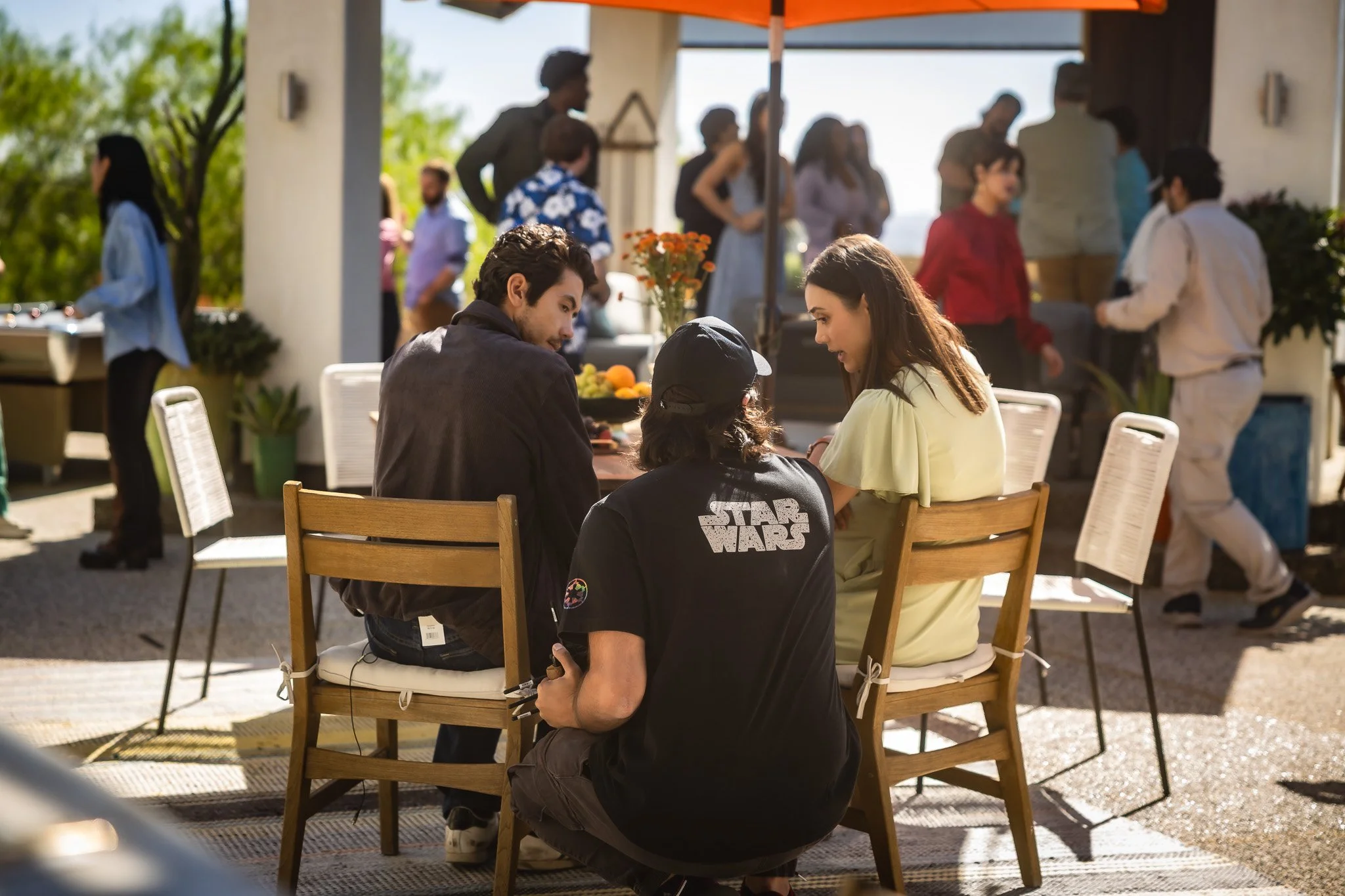 People sitting around a table engaging in conversation at an outdoor gathering or party, with others mingling in the background under a large orange umbrella.