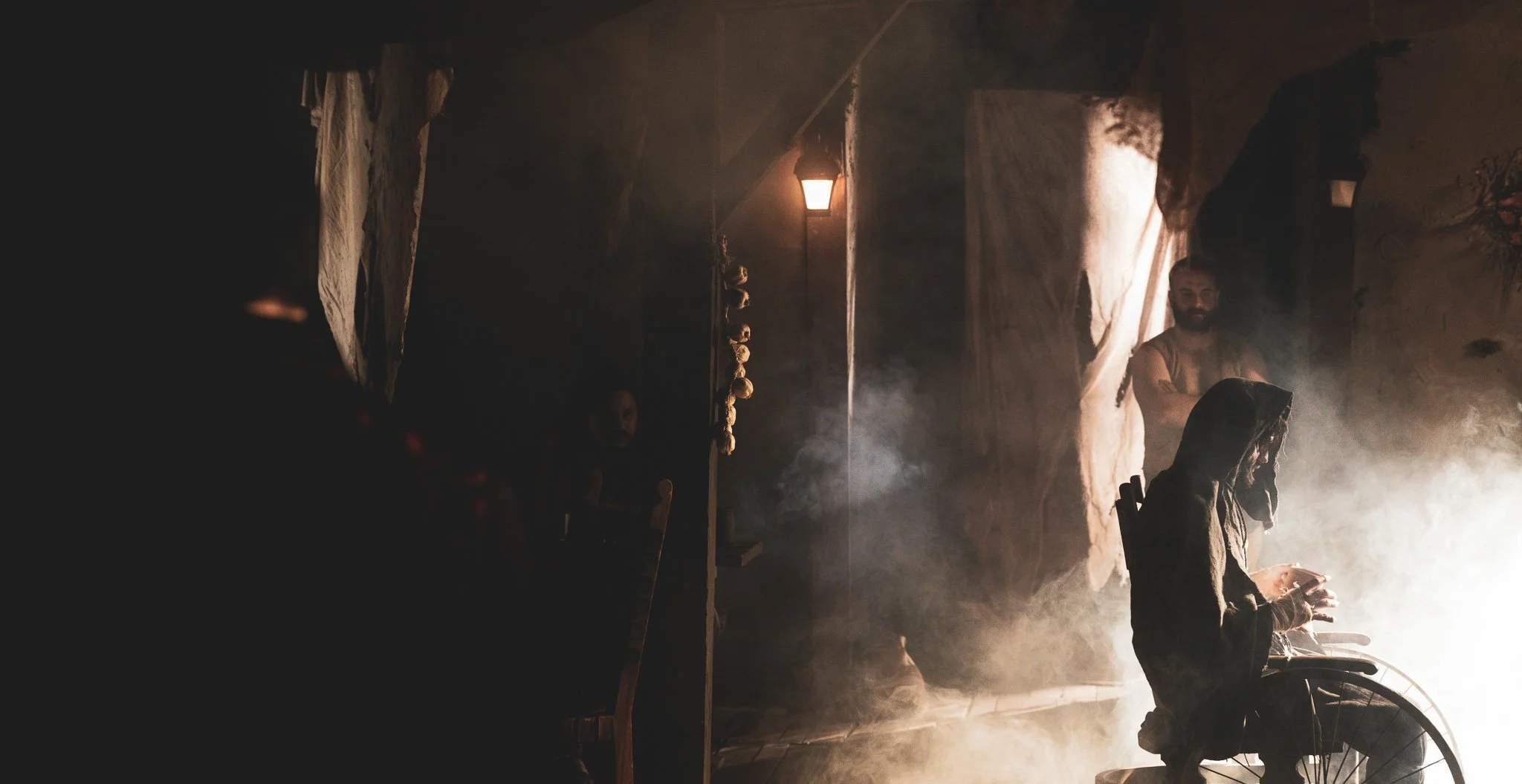 A person in dark clothing and hood sitting in a wheelchair, with their head lowered, in a smoky, dimly lit room with a person standing in the background, illuminated by a bright light source to the right.