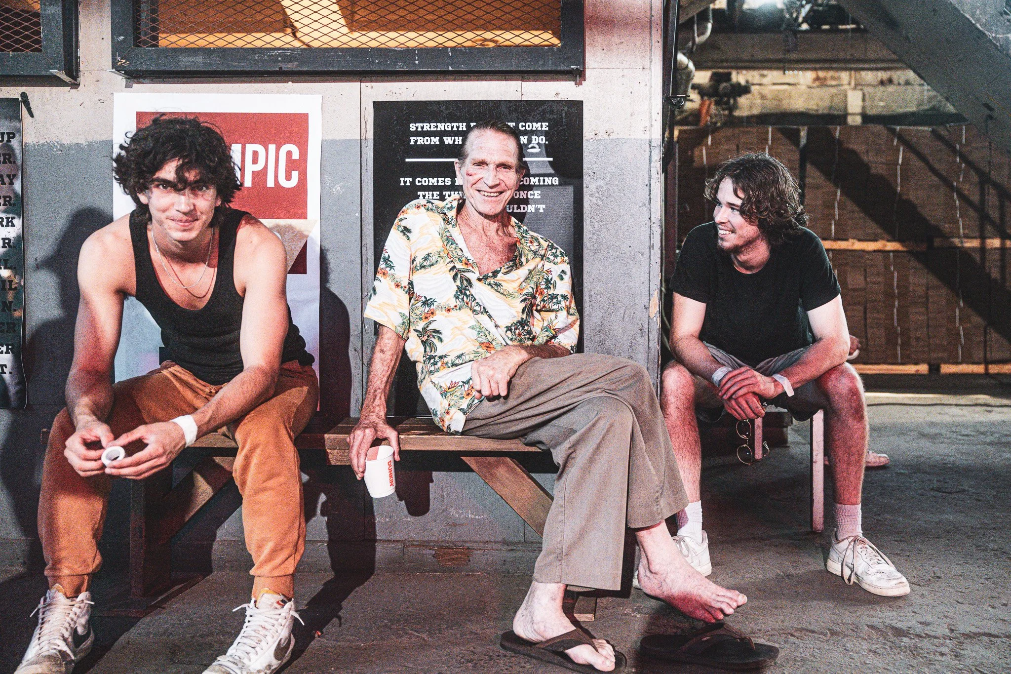 Three men sitting on a bench in an urban setting, with two posters on the wall behind them. The man in the middle is smiling and wearing a floral shirt, with one foot bare. The man on the left is wearing a black tank top and brown pants, and the man