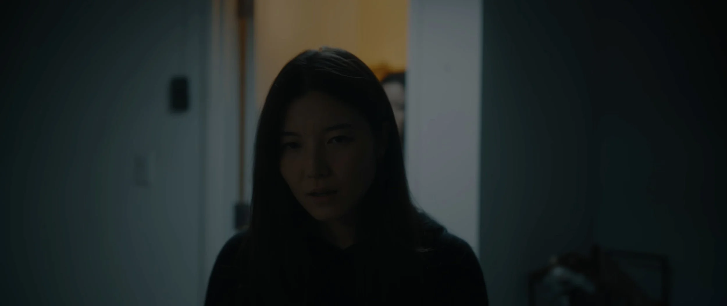 A woman with dark hair looking towards the camera in a dimly lit room with lockers in the background.