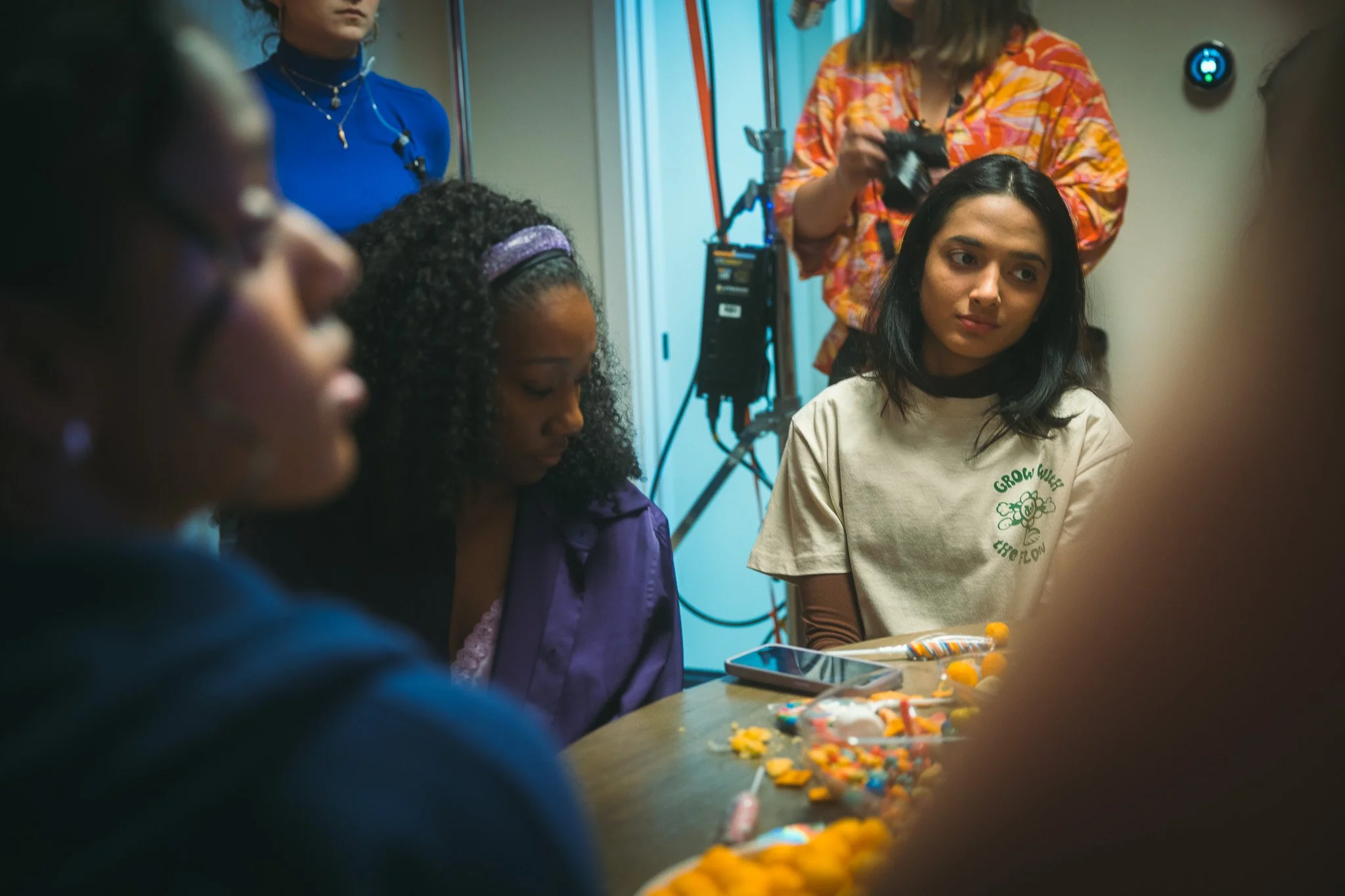 Group of young women sitting around a table with colorful cookies and snacks, in a room with a woman holding a camera and a camera tripod in the background.