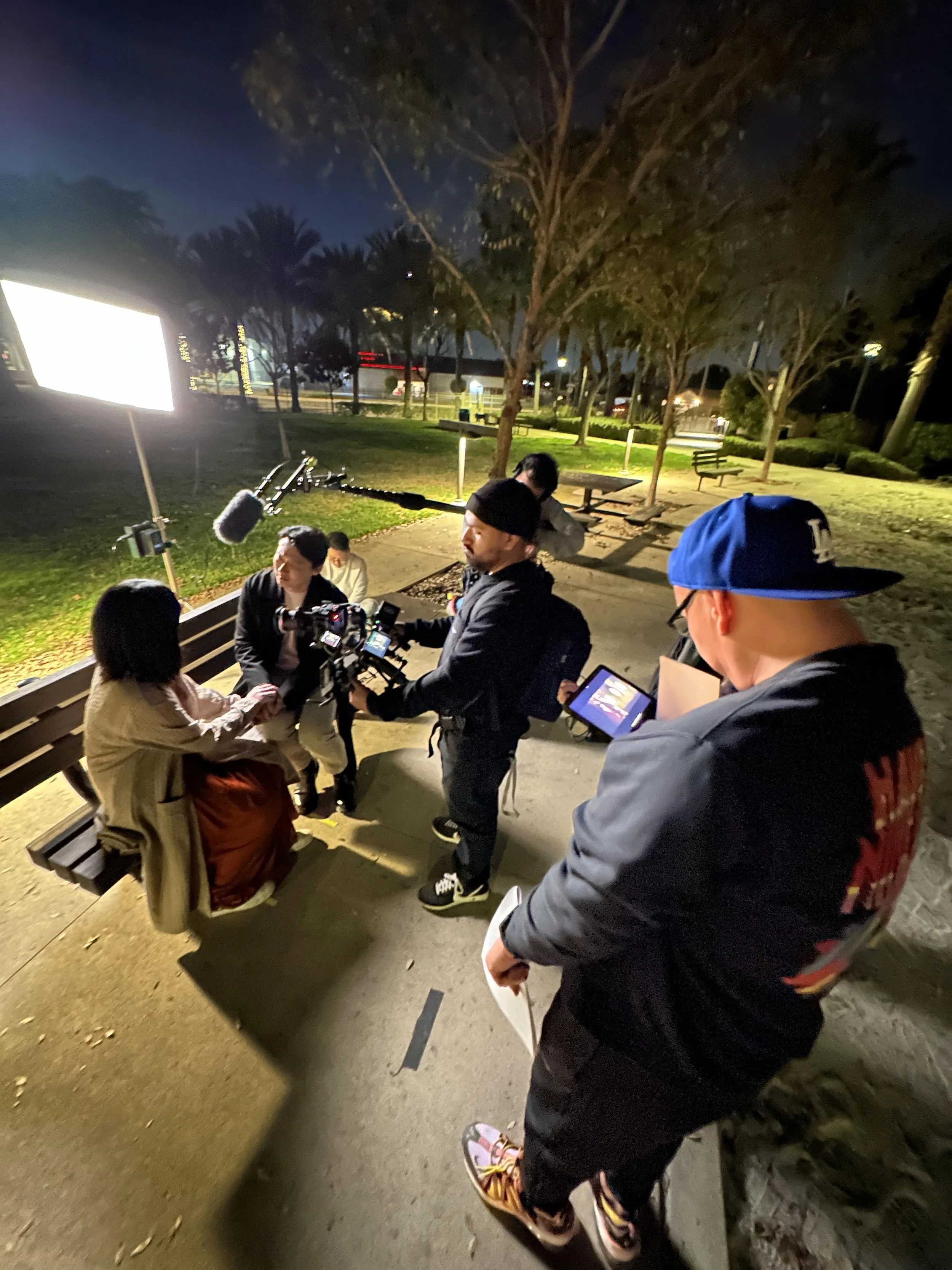 Group of people sitting and standing in a park at night, some with cameras and recording equipment, engaging in a conversation or interview.