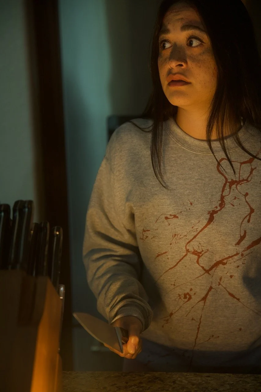 A woman with long dark hair and freckles, wearing a gray sweatshirt with bloodstain-like red splatters, holding a large kitchen knife in a dimly lit room, looks surprised or frightened.