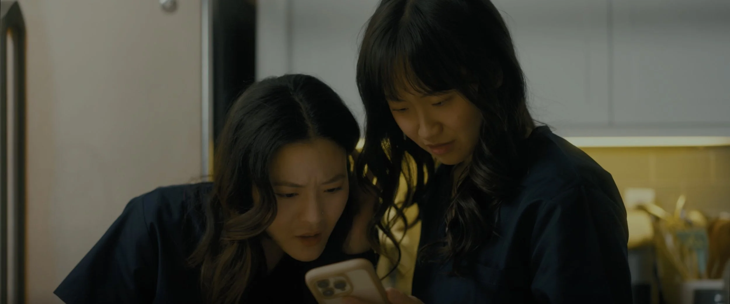 Two women with dark hair, one looking at a smartphone, the other leaning in closely, in a kitchen setting with yellow and gray cabinets.