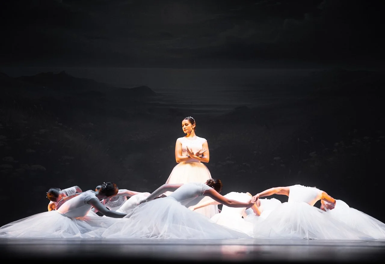 Ballet dancers perform on stage with a dark backdrop, one standing in a beige dress and five lying on the floor in white tutus, holding hands and leaning forward.