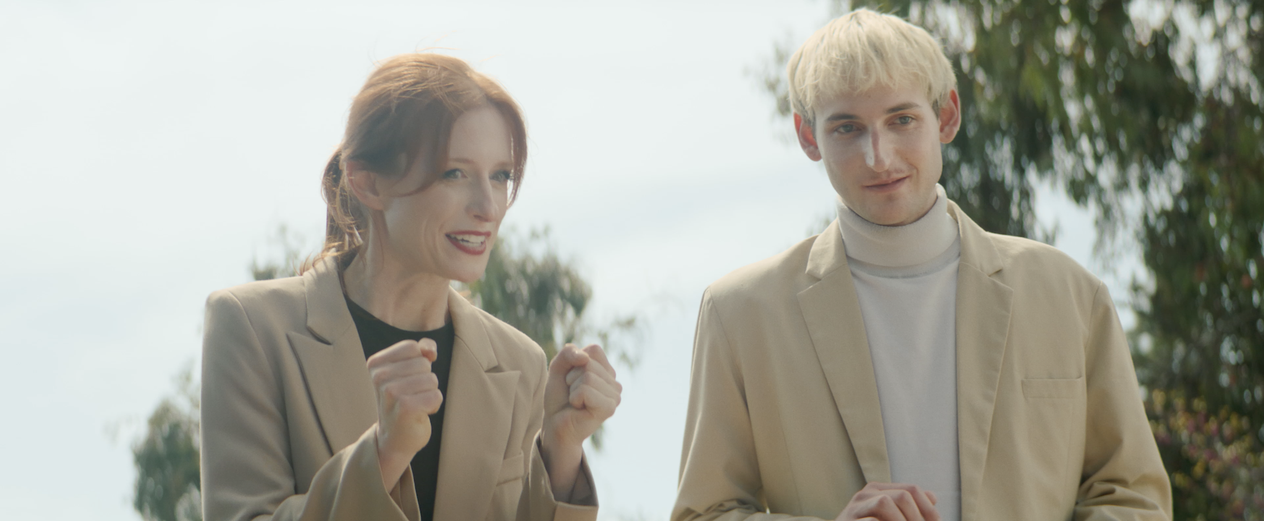 A woman with red hair smiling and fist-pumping next to a blond man smiling outdoors with trees in the background.