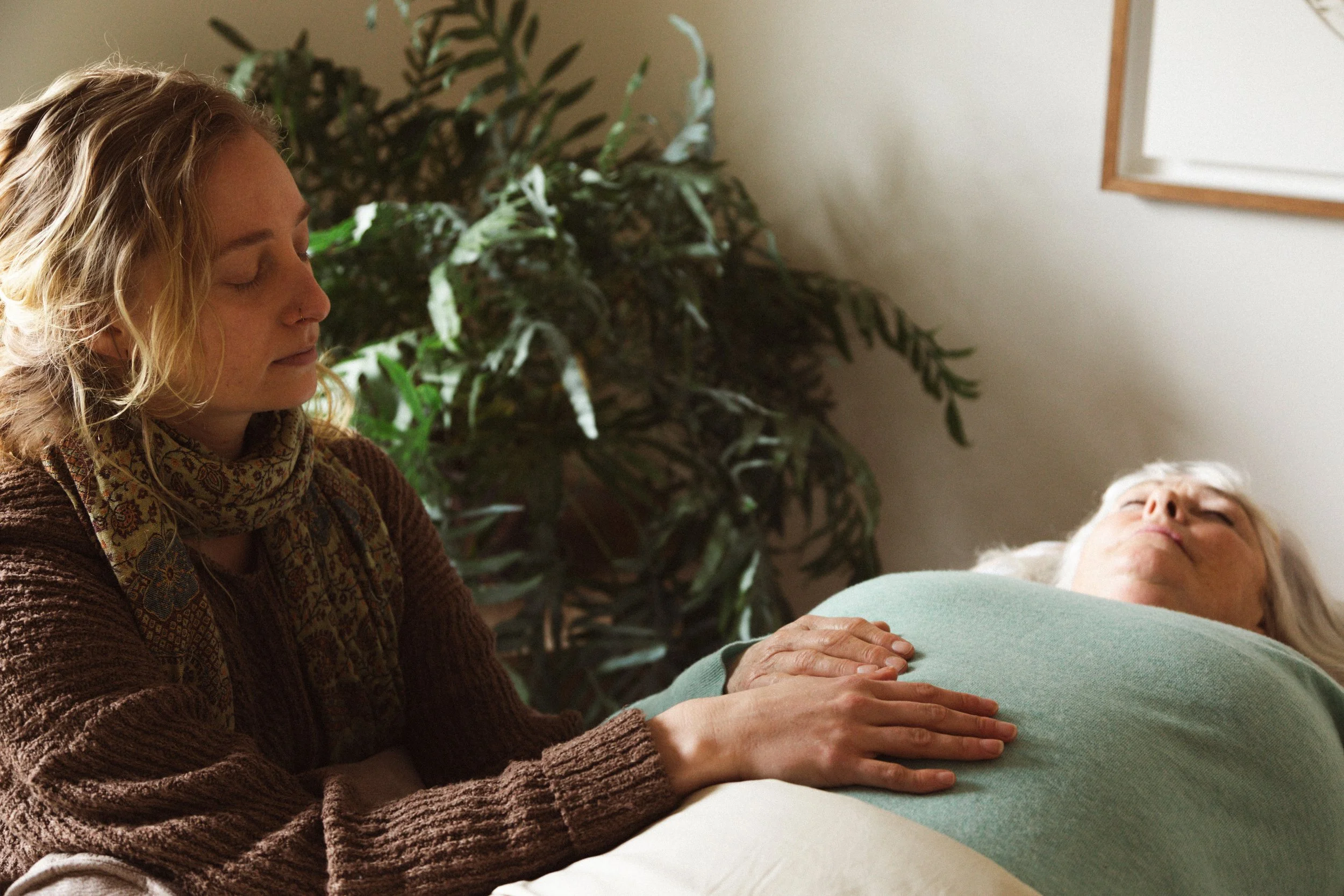 A woman with blonde, curly hair is sitting with her eyes closed, gently holding the hand of an elderly woman lying down, who has white hair and is wearing a green sweater. There is a large green plant in the background and a whiteboard on the wall.