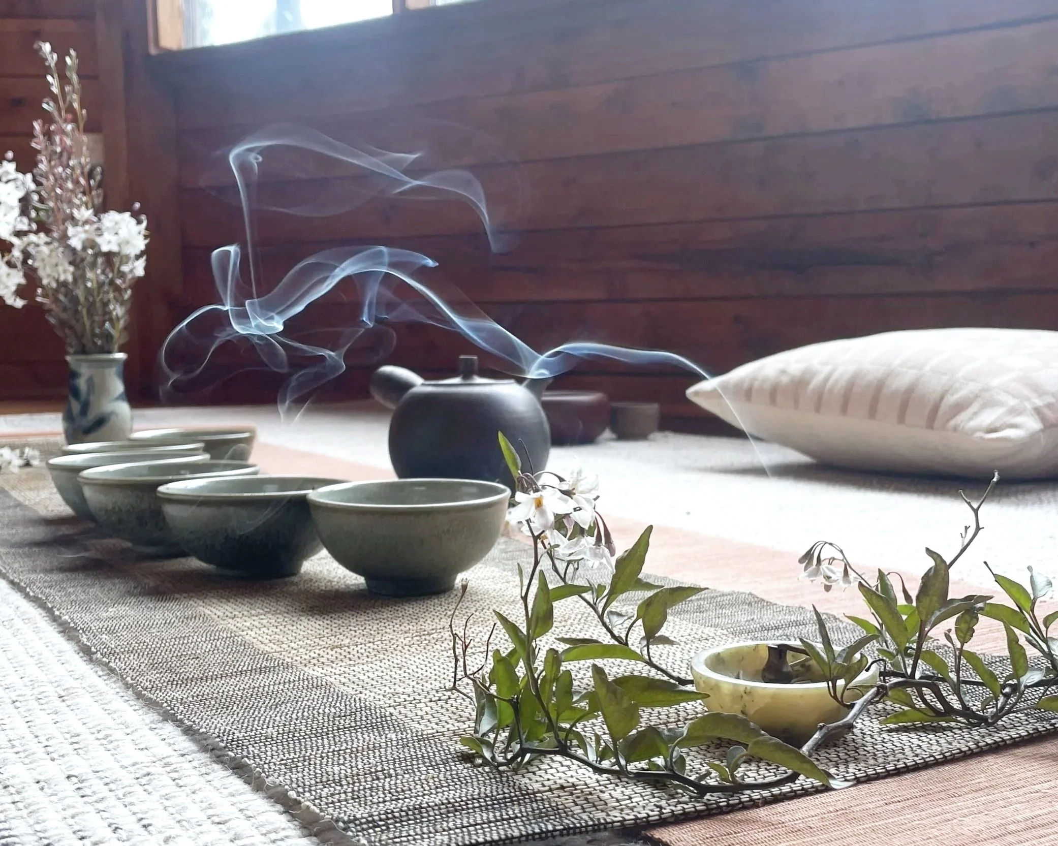 A cozy interior with a textured beige rug, a wooden wall, a blanket-covered bed, and a table with a teapot emitting steam, surrounded by cups, a vase of white flowers, and some small potted plants.