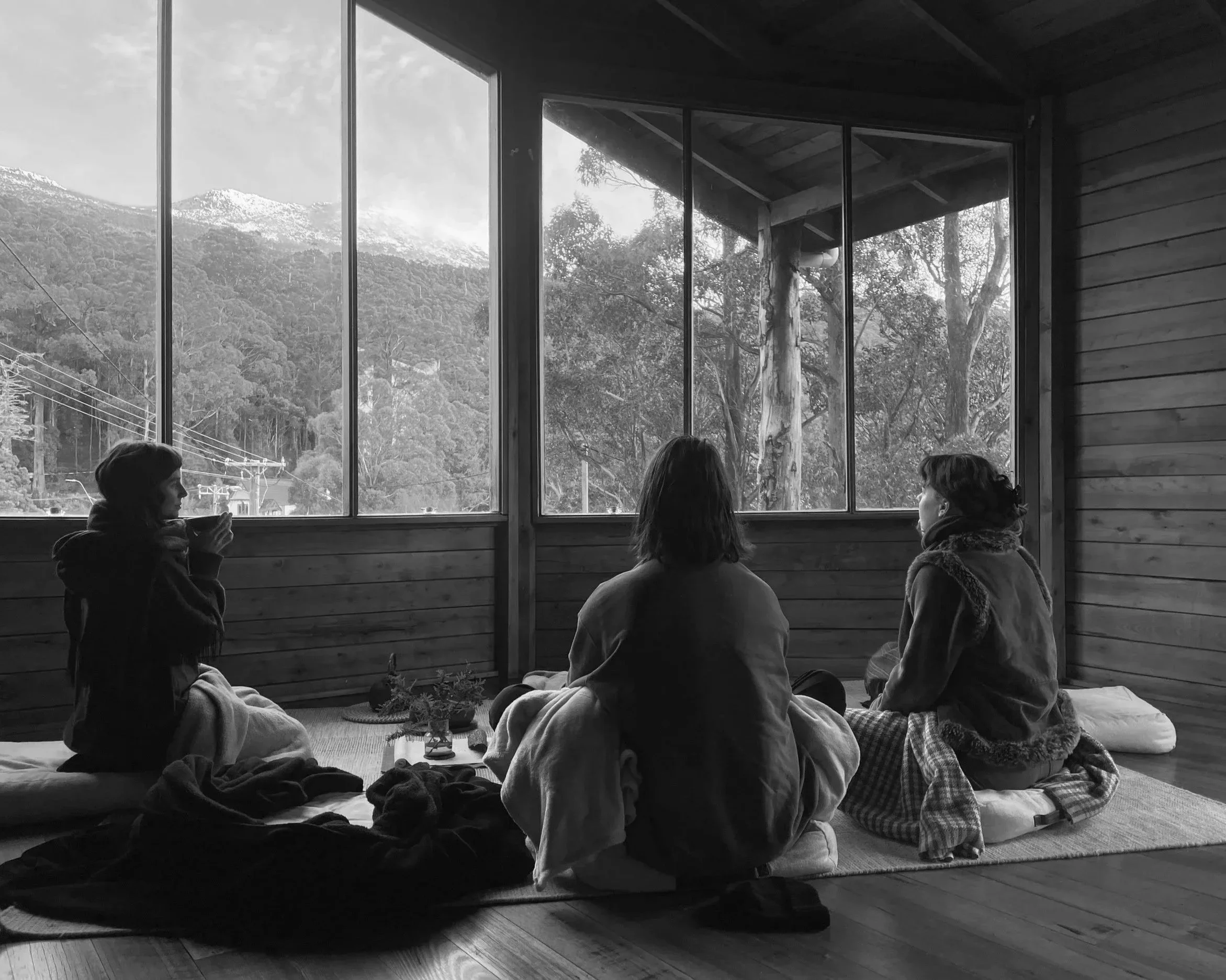 People sitting cross-legged on the floor inside a room with large windows showing mountain scenery outside, possibly participating in a meditation or yoga session.