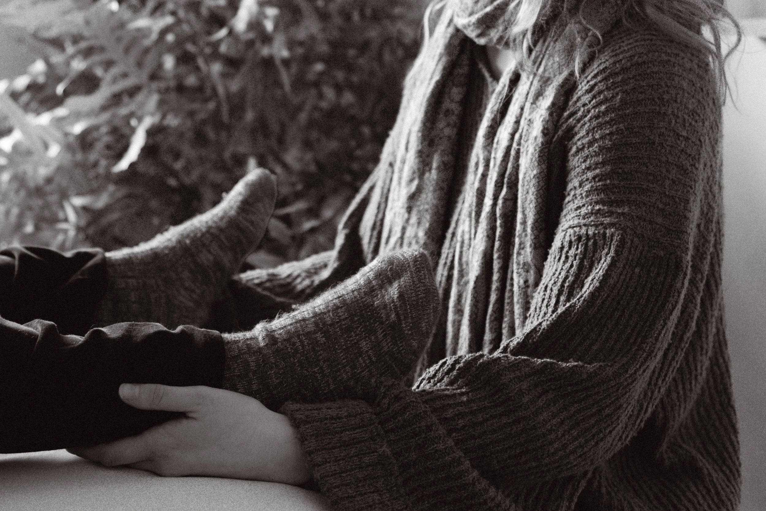 Close-up black and white photo of two people holding hands, with one person touching the other's arm. One person is wearing a textured sweater, and the other is wearing a dark long-sleeve shirt, with their arms resting on a flat surface.