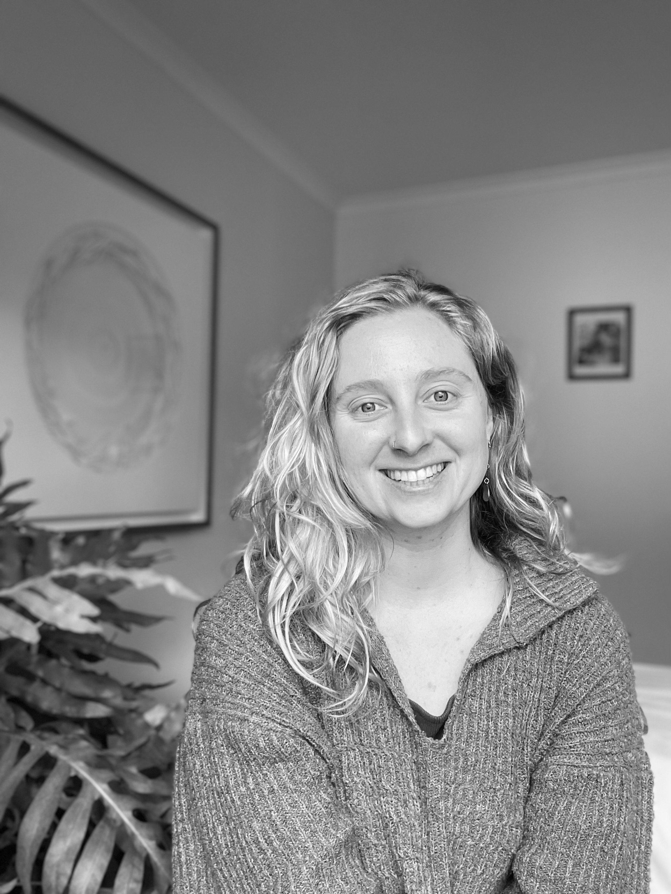 Smiling woman with blonde wavy hair wearing a textured sweater, indoors with framed pictures and a plant in the background.