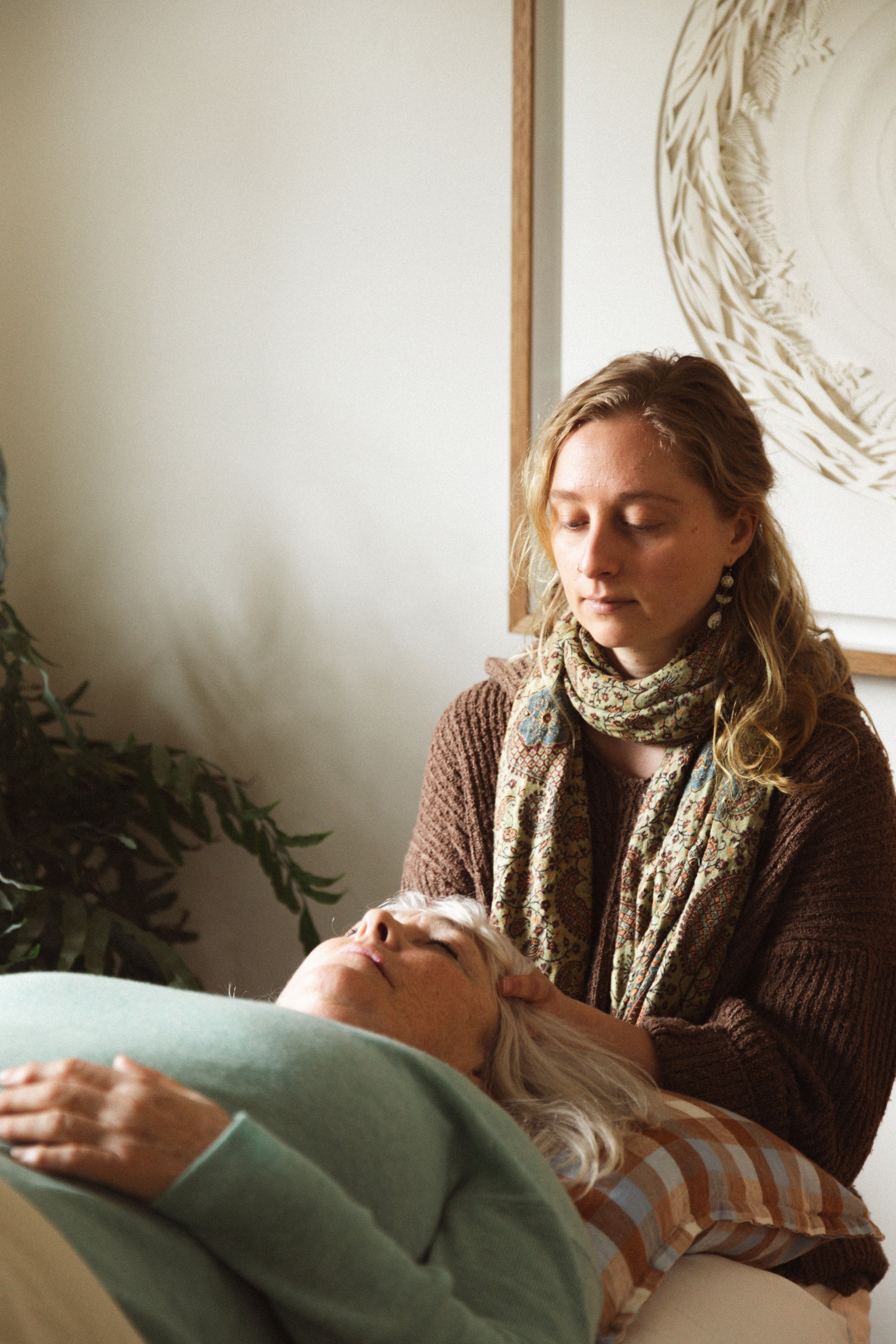 A woman providing care or massage to an elderly person lying down on a bed or couch in a cozy room, with a plant and wall art in the background.