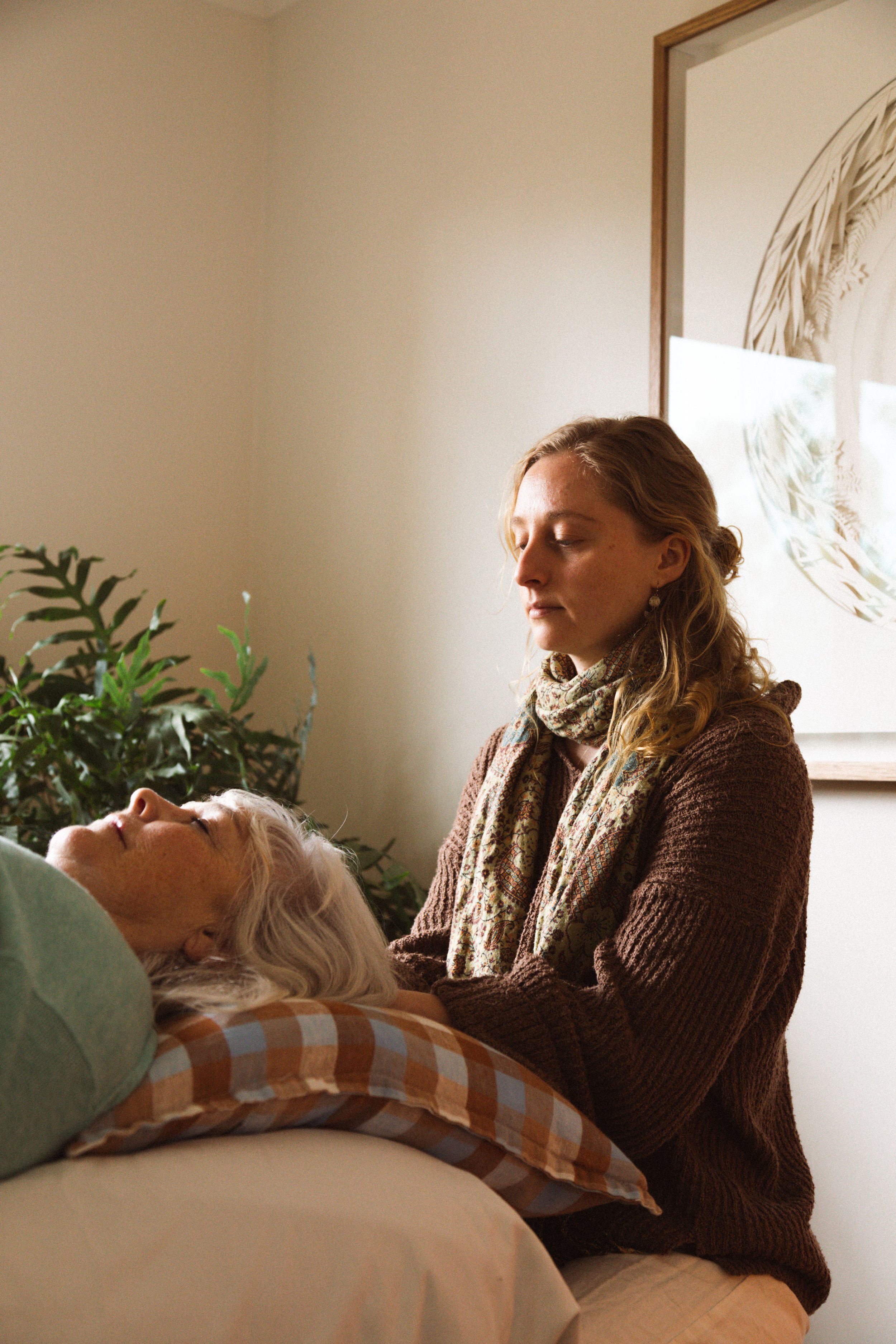 A woman with long, curly gray hair lying in bed with her eyes closed, while a younger woman with long, straight hair and earrings sits nearby, looking at her with a calm expression. There are plants and framed artwork on the beige wall behind them.