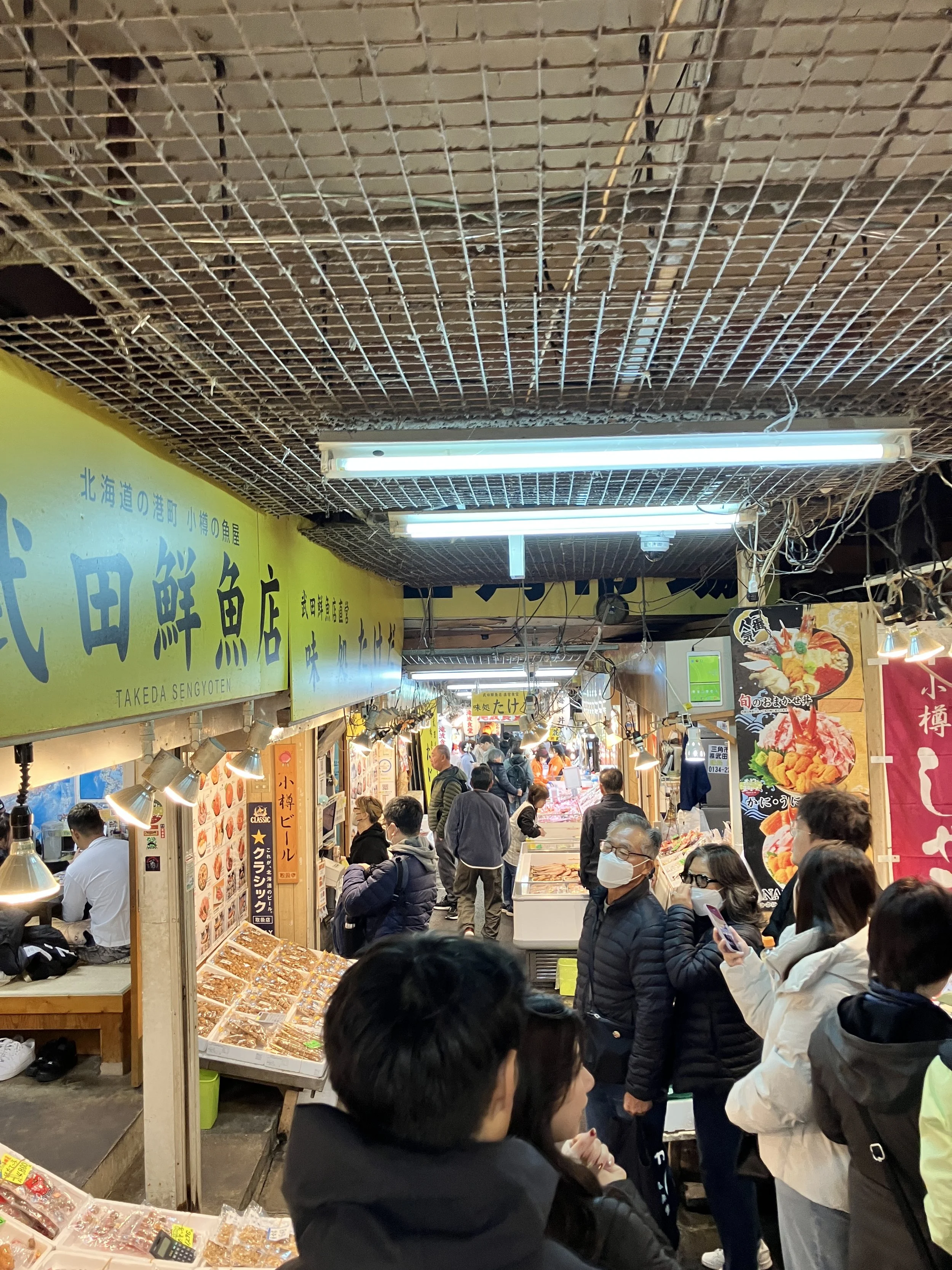 Crowded indoor market with people shopping and dining under fluorescent lights, various food stalls, and signs in Japanese.
