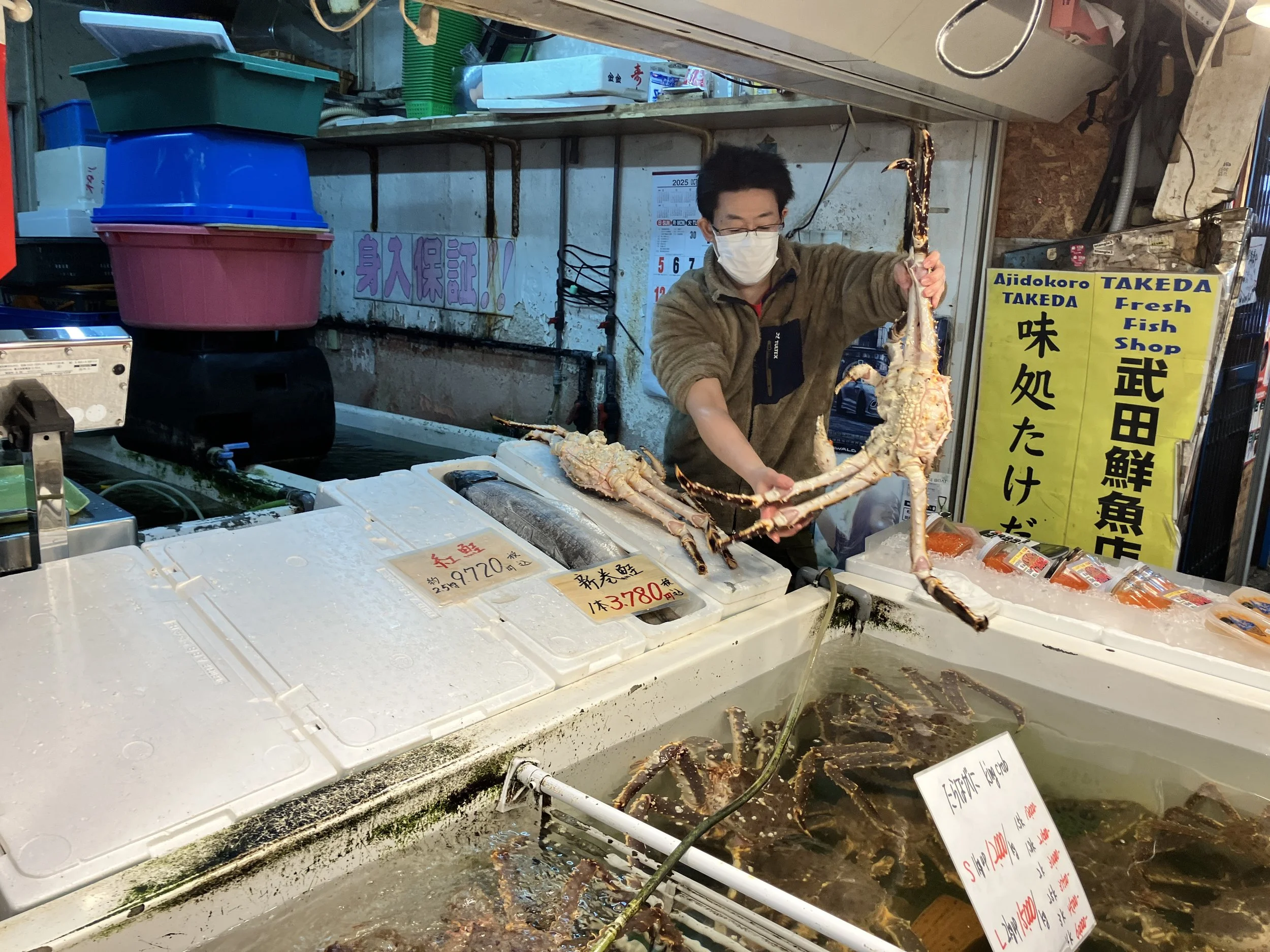 A man wearing a mask and glasses is holding a large crab at a seafood market stall with various fresh seafood items on display.