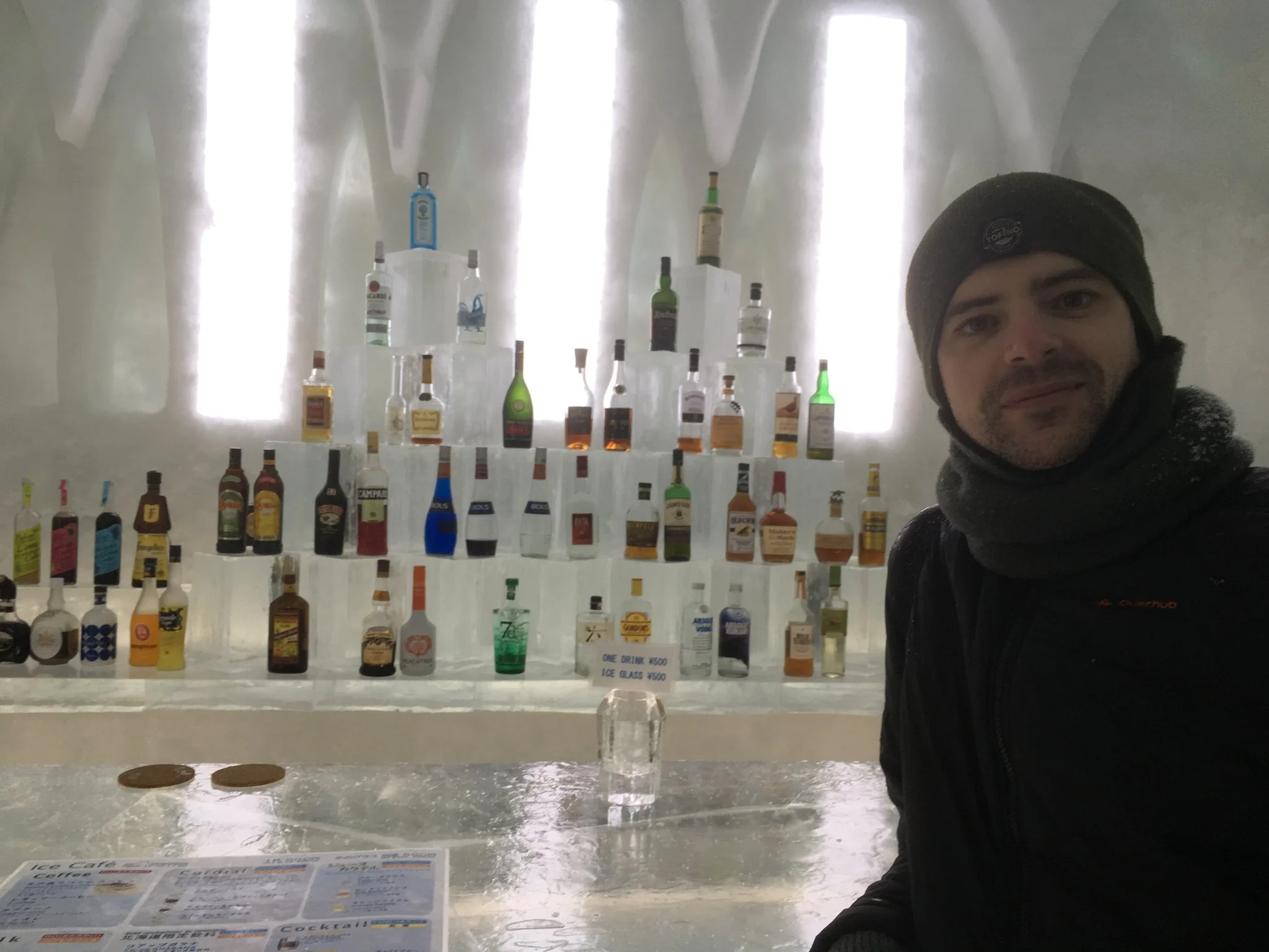 A man wearing a black beanie, black jacket, and scarf sitting in front of a display of ice sculptures and bottles of liquor inside a cold ice room or bar.