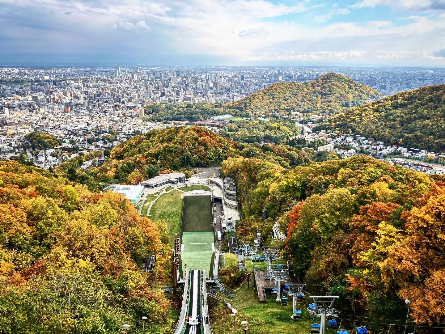 Aerial view of a ski jump overlooking a city with a forested hill, a mountain, and a cloudy sky.