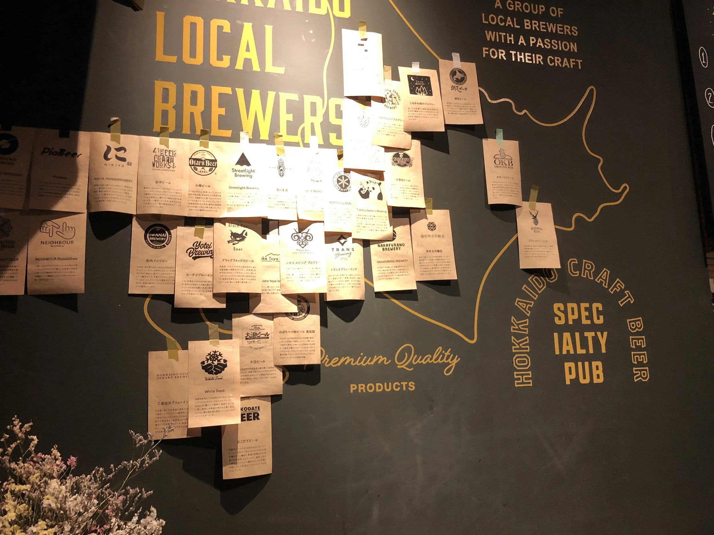 Mapa y lista de cervecerías artesanales locales en un cartel de pub en Sapporo, decorado con notas adhesivas y flores en la esquina inferior izquierda.