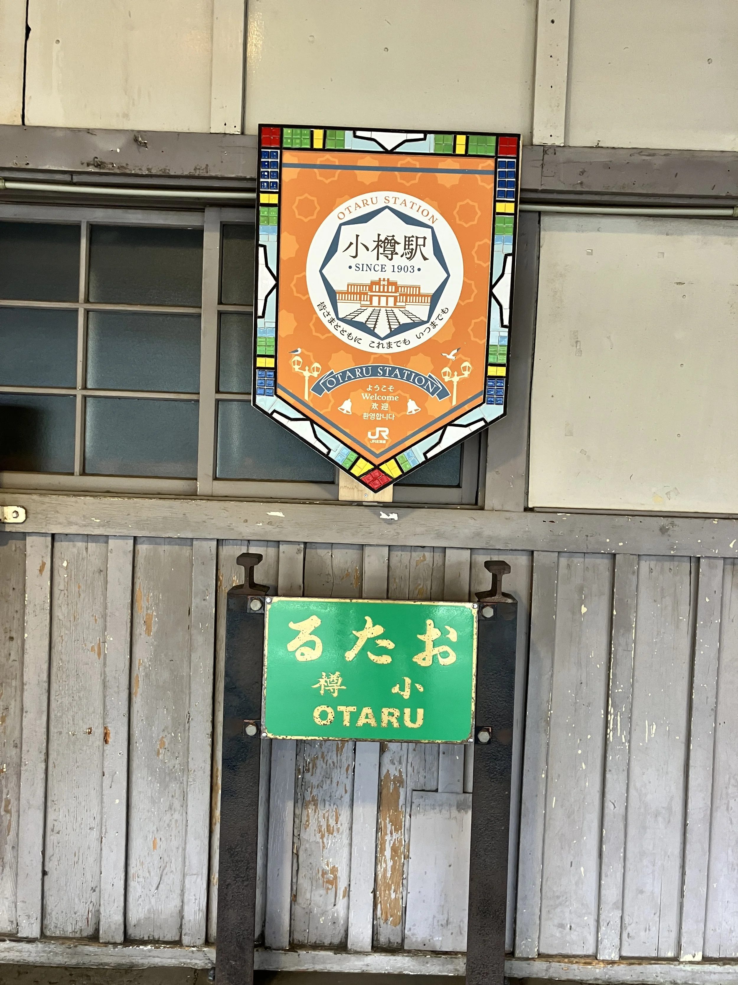 A colorful sign with a decorative border above a green sign on a wooden wall, indicating Otaru Station with Japanese characters and a small bilingual sign welcoming visitors.