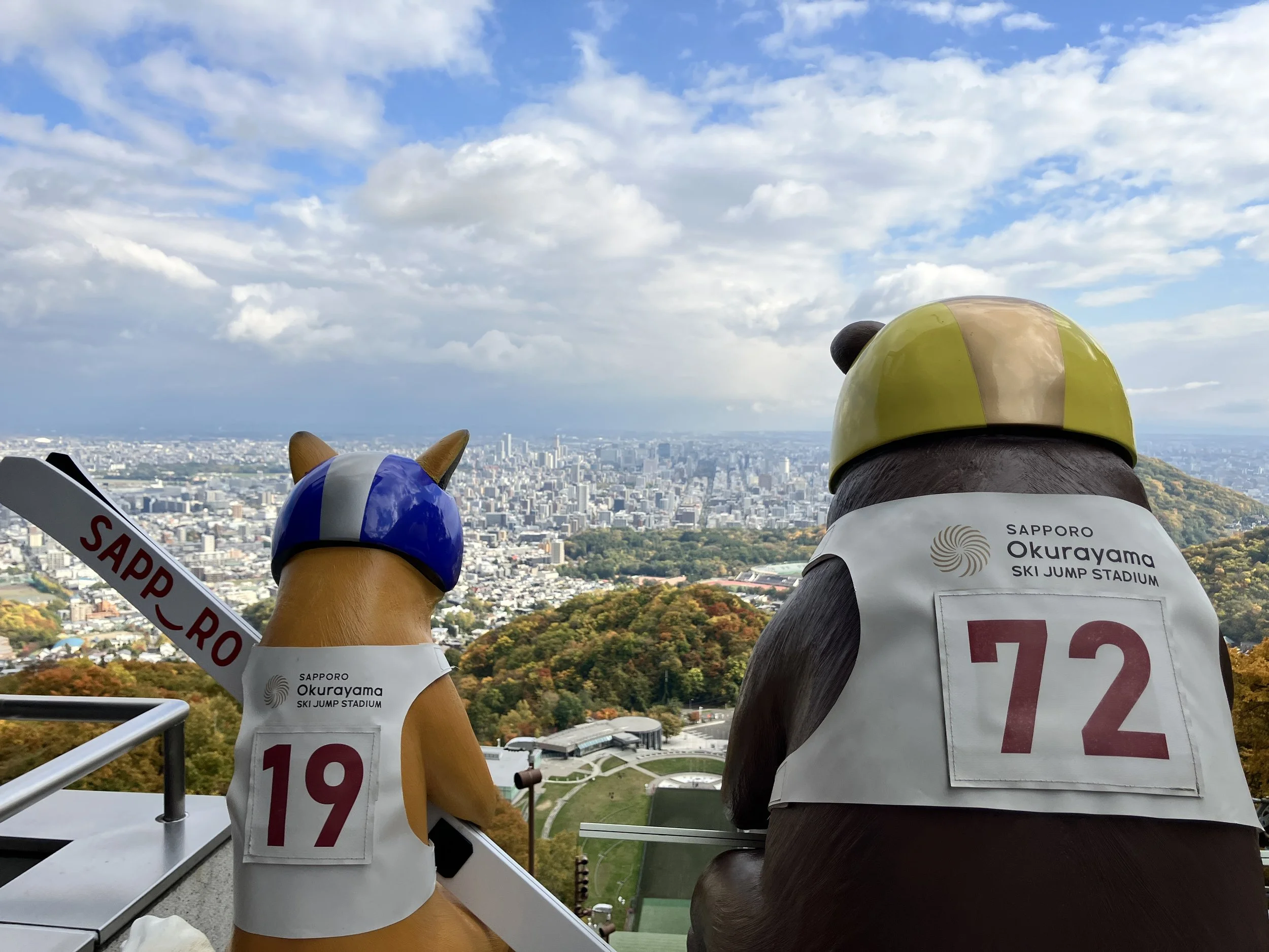 Sculptures of a dog and a bear wearing helmets and racing vests with numbers, overlooking the Sapporo cityscape from Okurayama ski jump stadium in Japan.