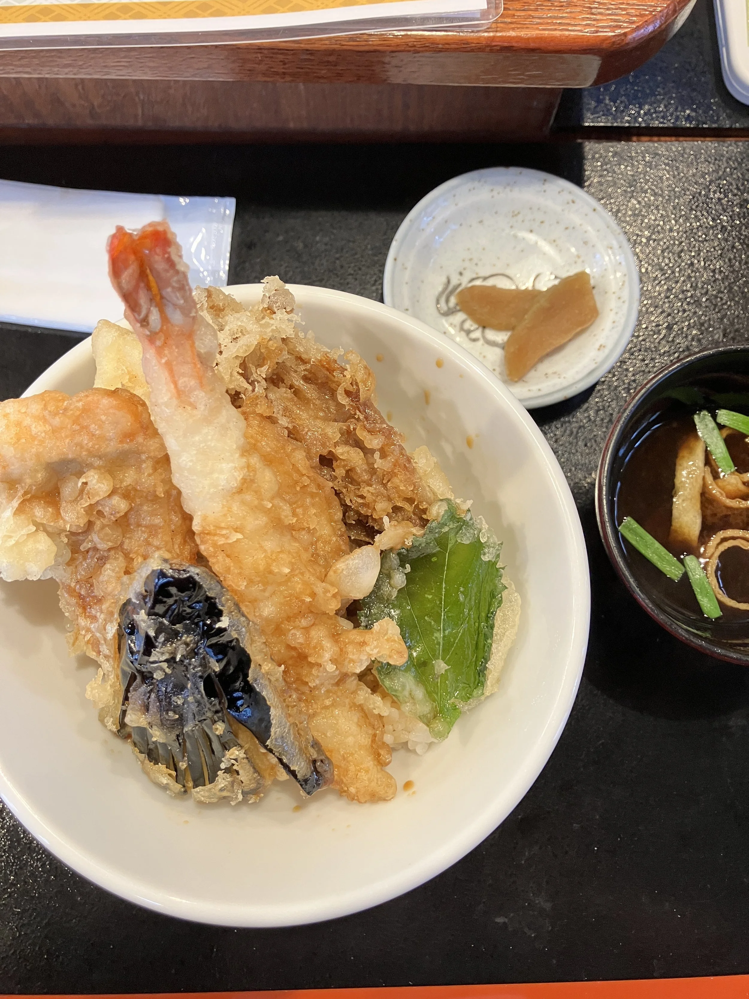 A bowl of assorted tempura including shrimp, eggplant, and greens, with a small dish of dipping sauce, a bowl of miso soup with green onions, and a small dish of pickled ginger on a black tray.