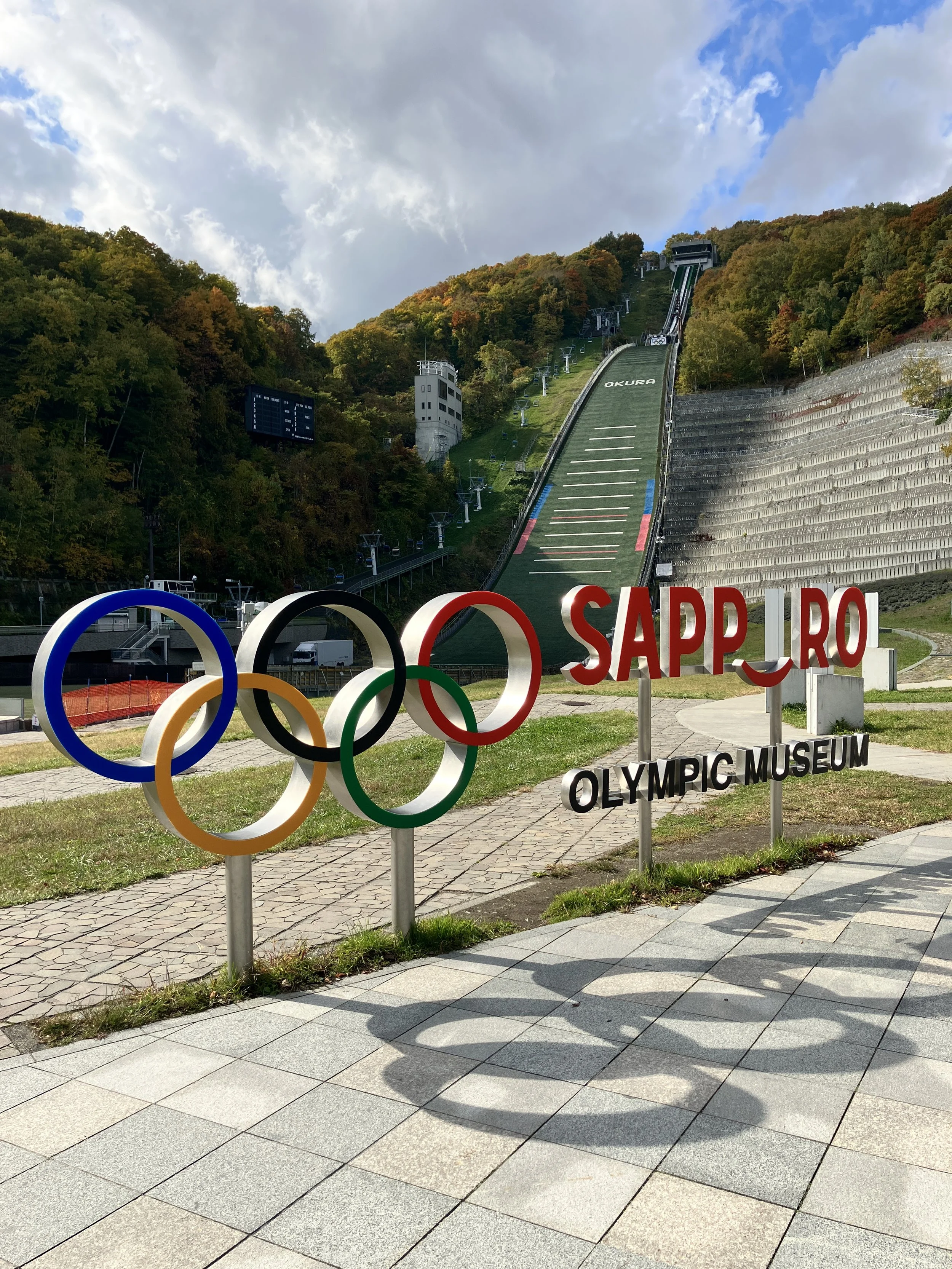 Sapporo Olympic Museum sign with Olympic rings and ski jump in the background amidst trees and cloudy sky.