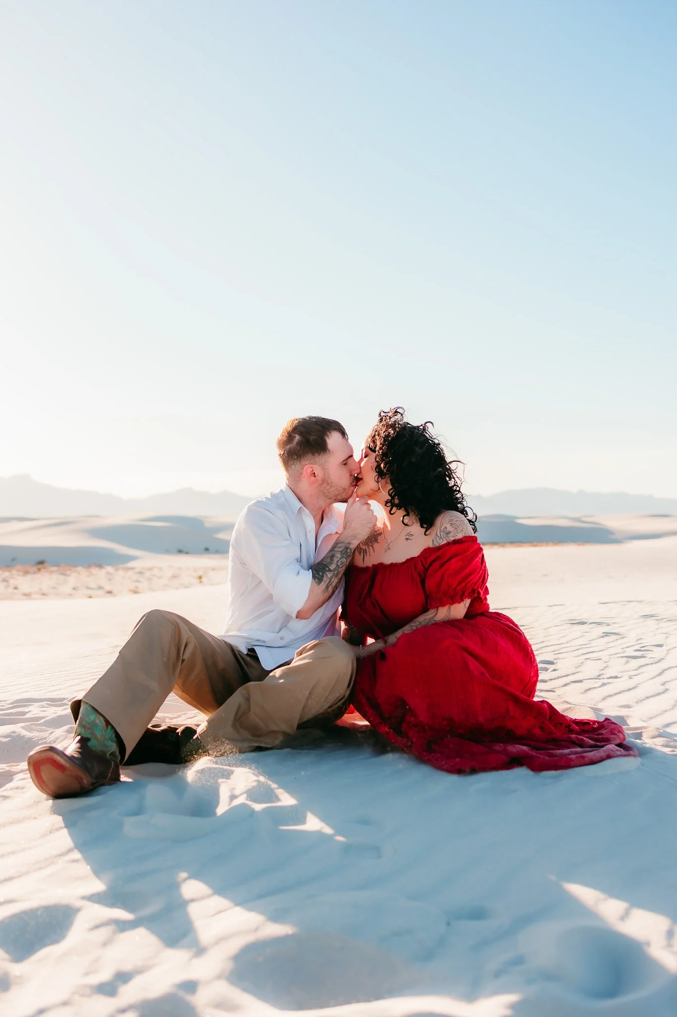 Couple sharing a kiss while sitting together on the dunes during their White Sands National Park engagement photography session.