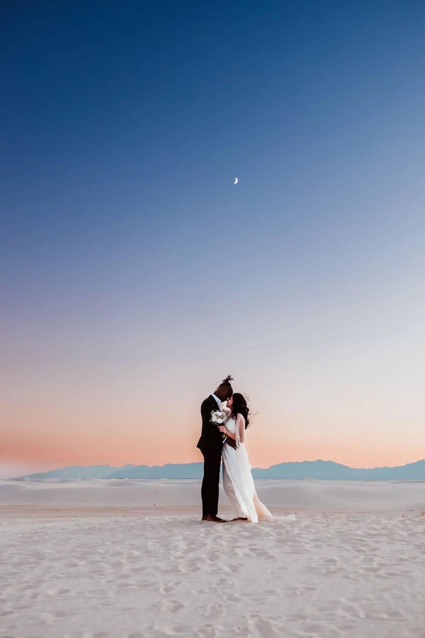 Bride and groom kissing beneath the evening sky and crescent moon after their White Sands National Park elopement in New Mexico.