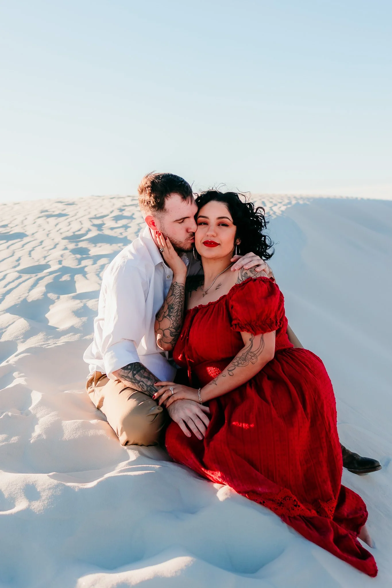 Engaged couple cuddling together on the white sand dunes during a sunset engagement session at White Sands National Park in New Mexico.