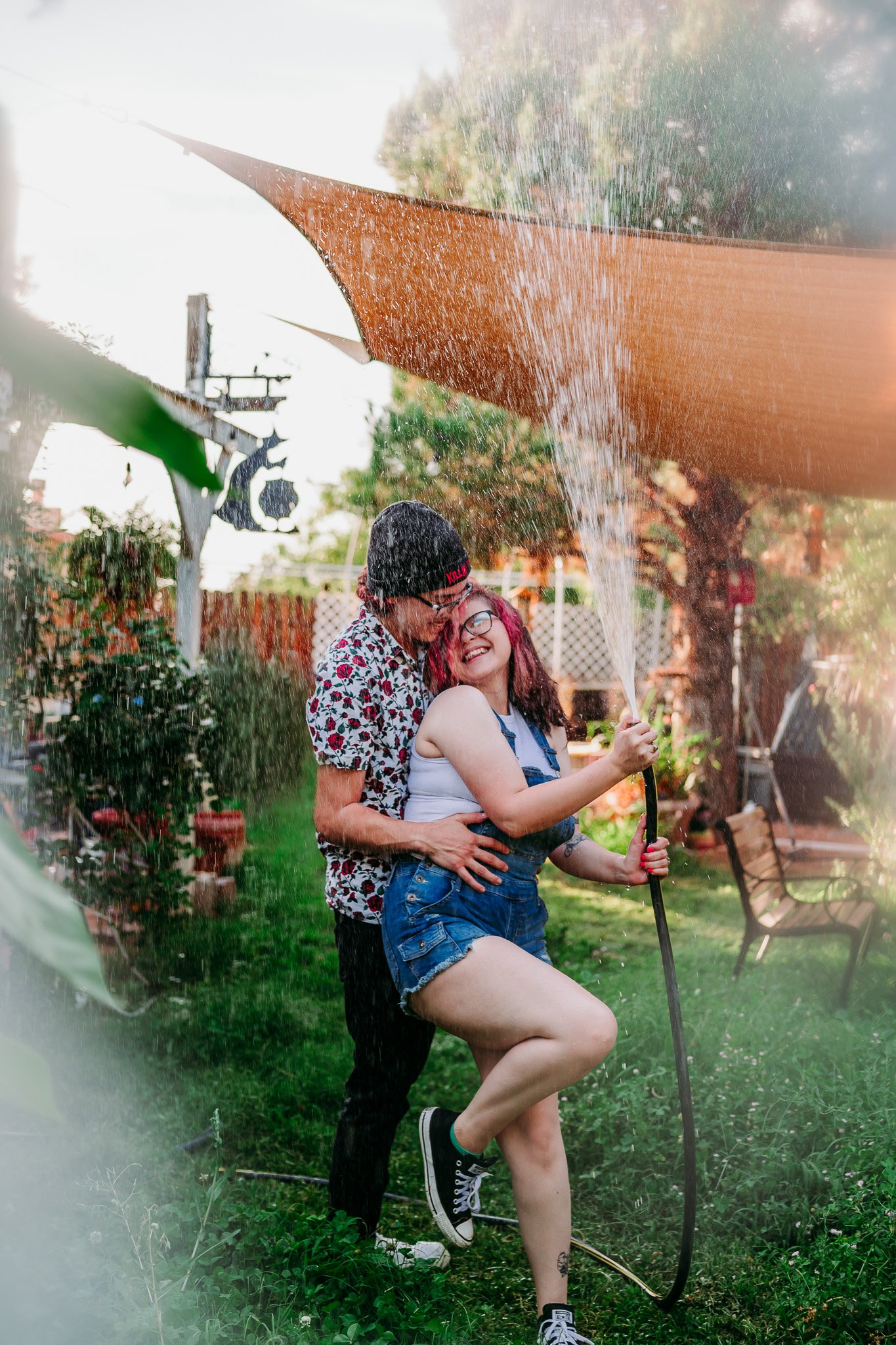 A young couple holding eachother in a backyard garden playing with the water hose & spraying into the air in a playful way. las cruces engagement photographer