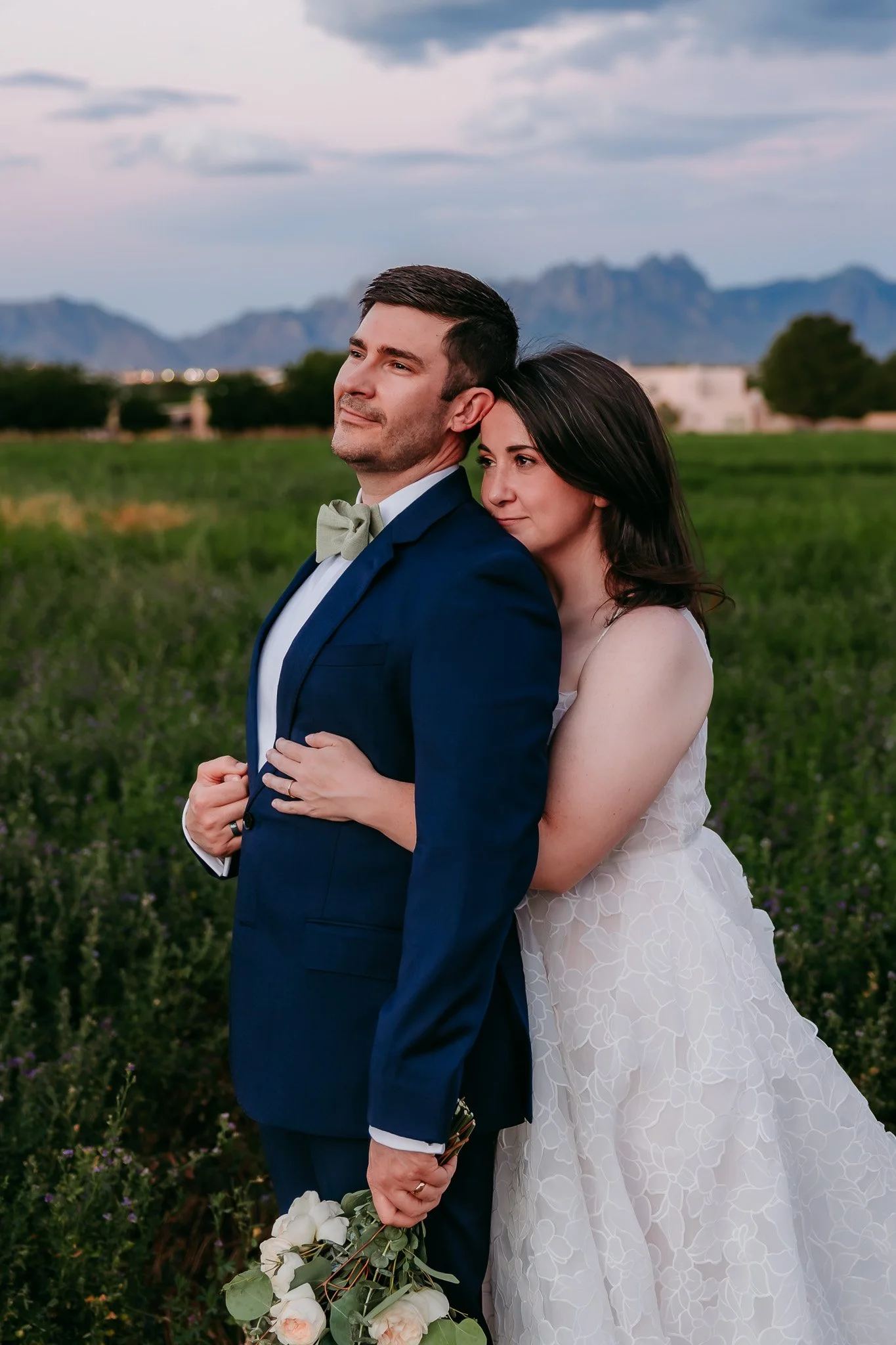 Bride hugging her groom from behind in an alfalfa field at sunset with the Organ Mountains in the background near Las Cruces, New Mexico.