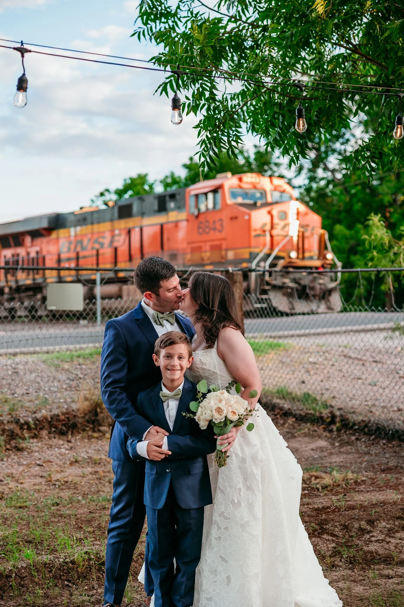 Bride, groom, and their son standing together in front of a passing train during their wedding elopement in the Mesilla Valley near Las Cruces, New Mexico.