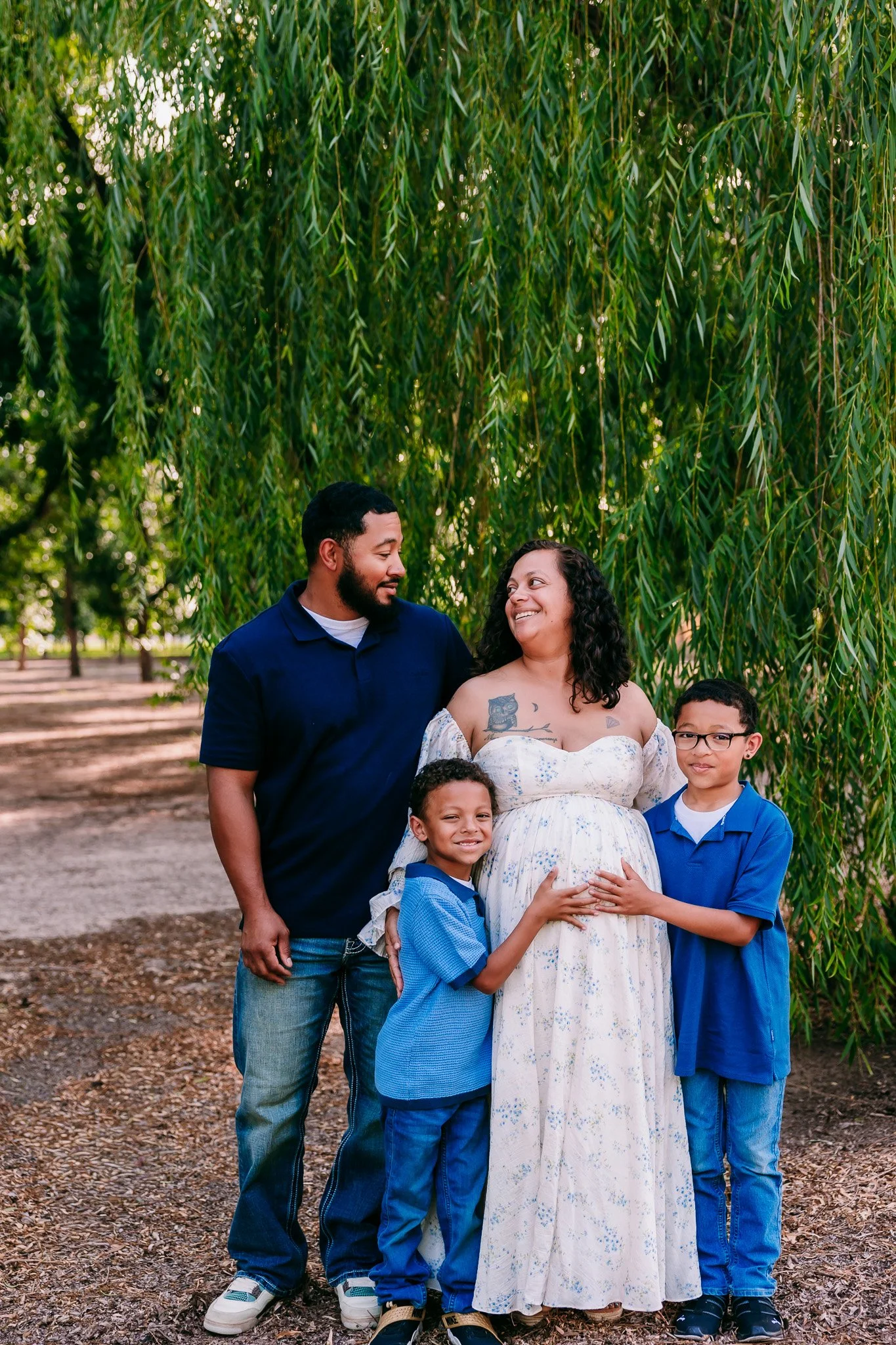 Family of four celebrating their pregnancy beneath willow trees during a maternity photography session in Las Cruces, New Mexico