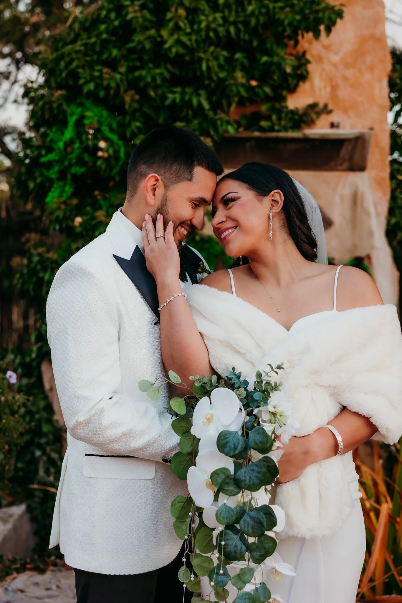 A bride and groom smile and touch foreheads outdoors, with the bride holding a cascading bouquet of greenery and white flowers, during a wedding celebration.