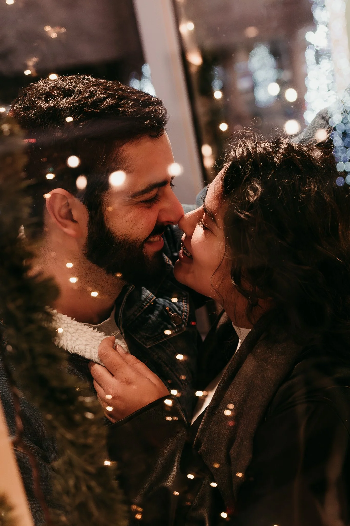 A couple with smiles face to face close to each other near a window with reflections of holiday lights, surrounded by blurred Christmas decorations.