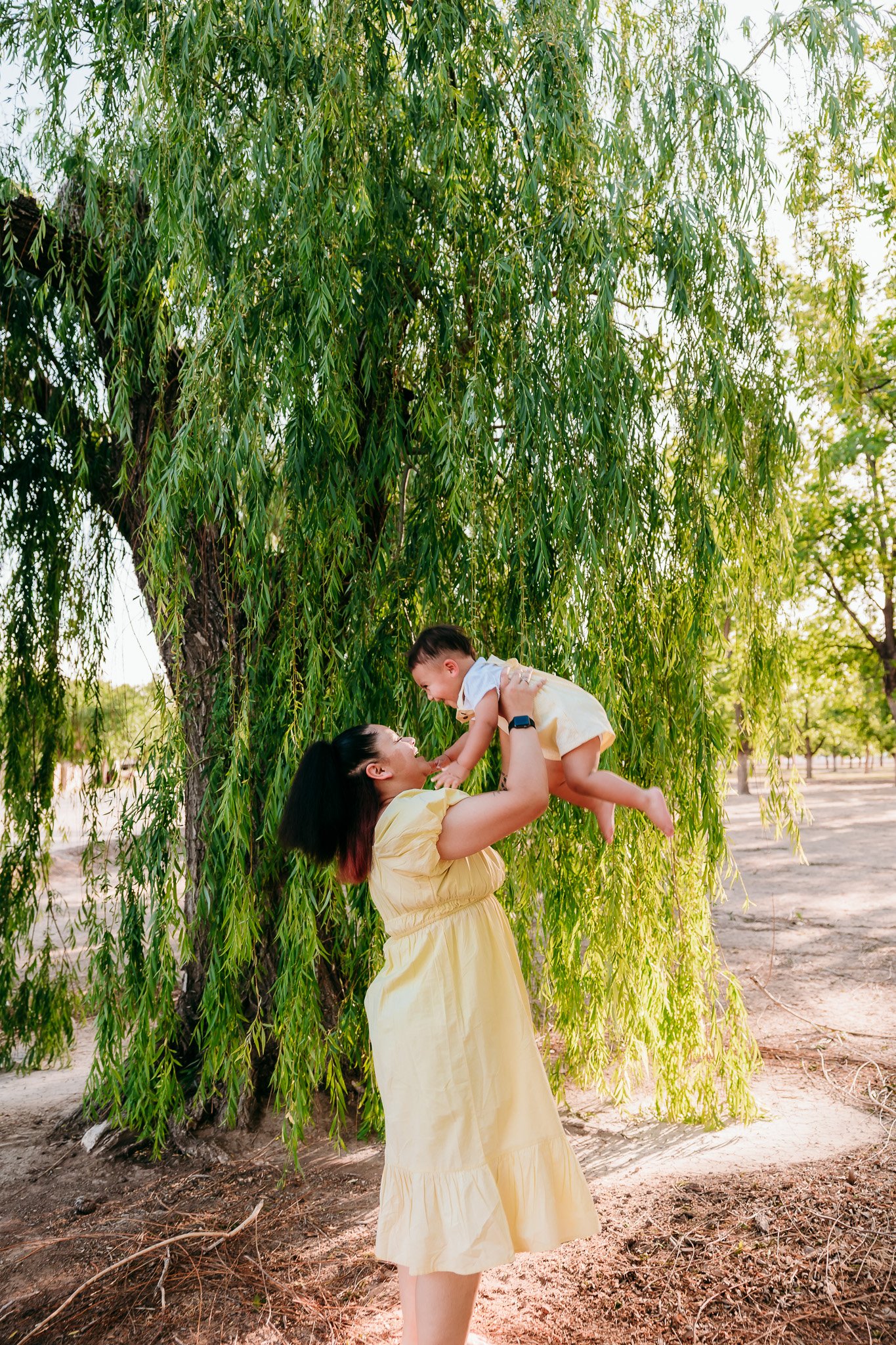 Mother lifting her child into the air beneath willow trees during a joyful outdoor family photography session in Las Cruces.
