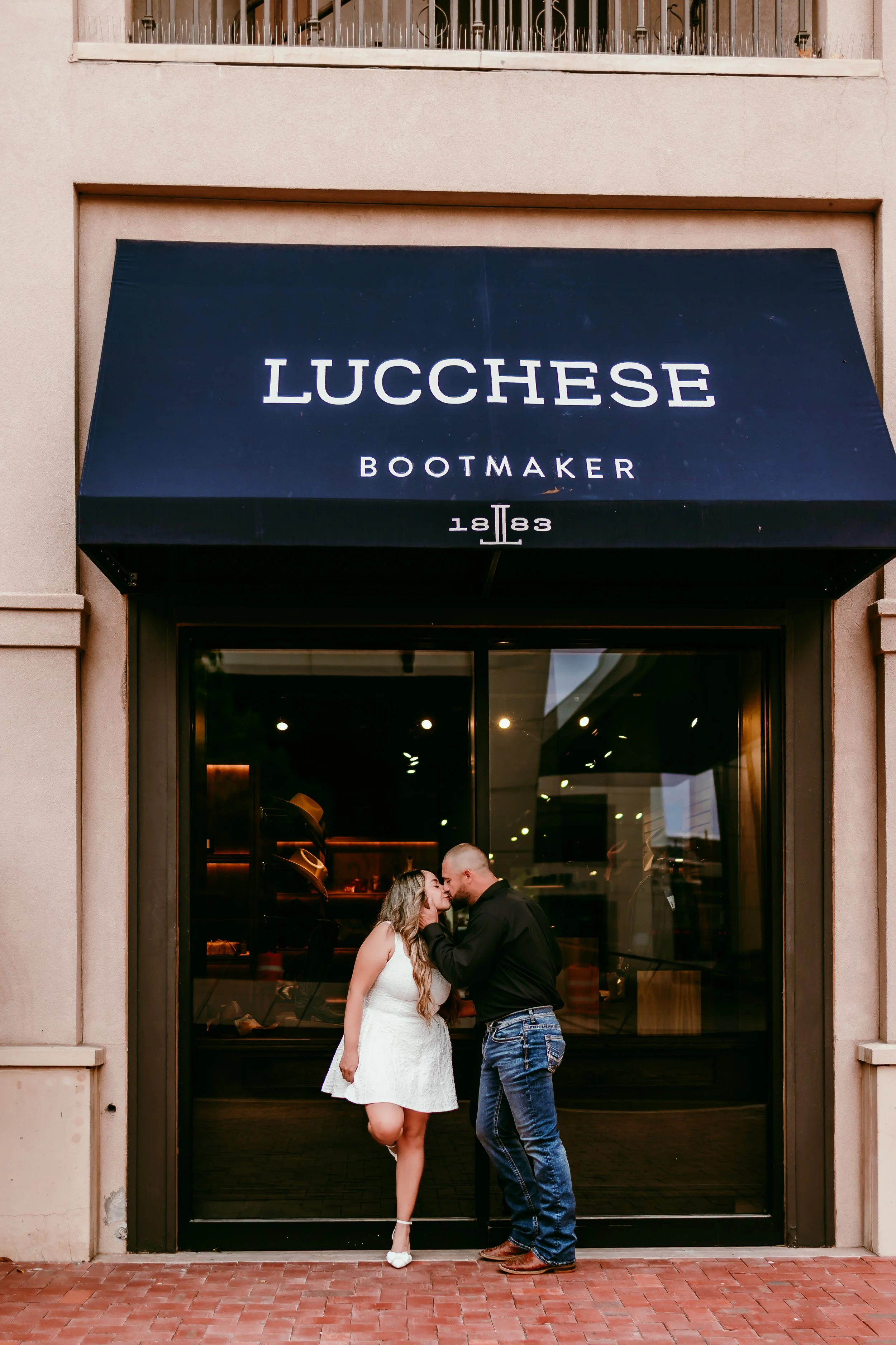 Engaged couple sharing a kiss outside Lucchese Bootmaker during a downtown El Paso engagement session.