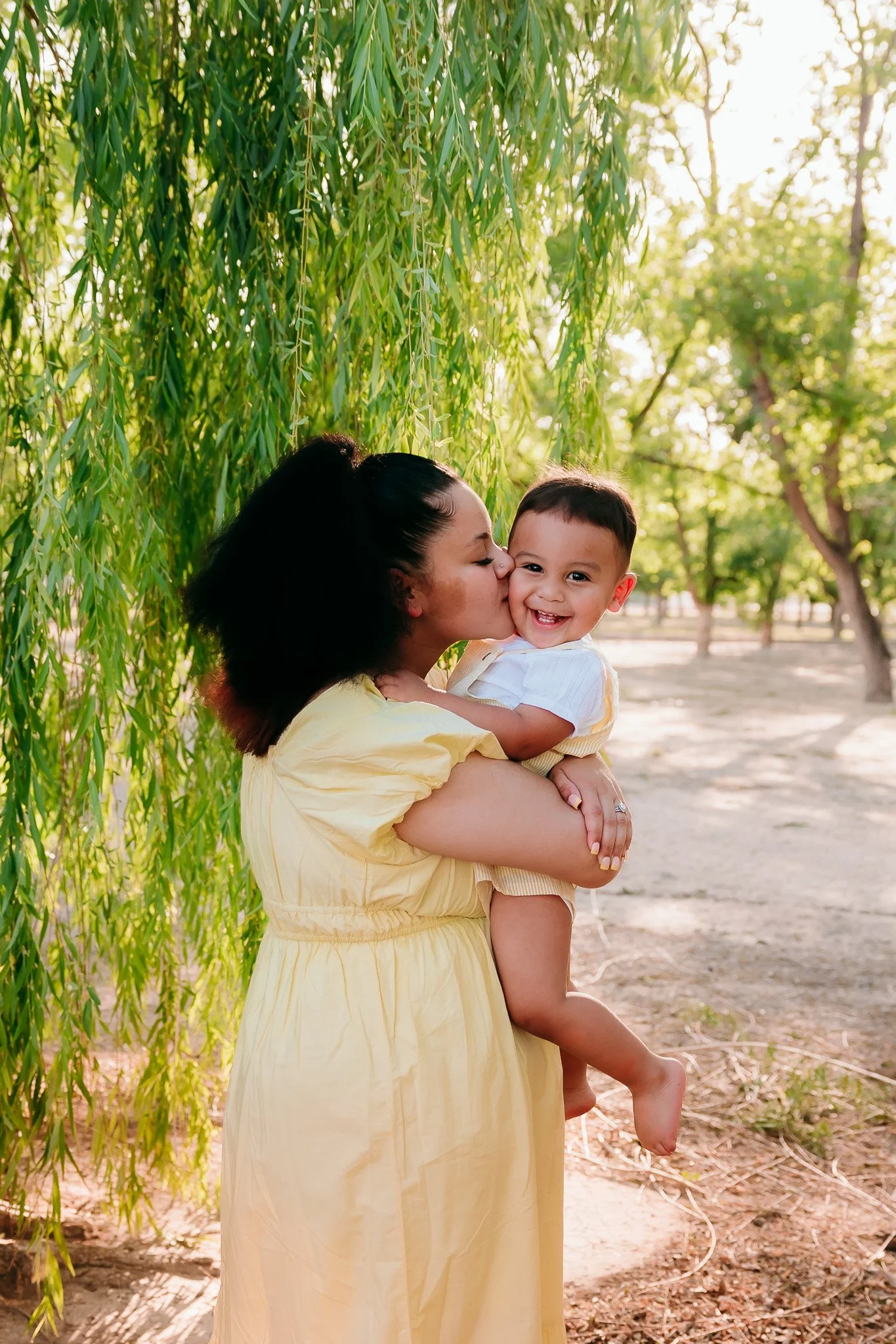Mother holding her toddler during a candid family photography session beneath willow trees in Las Cruces, New Mexico.