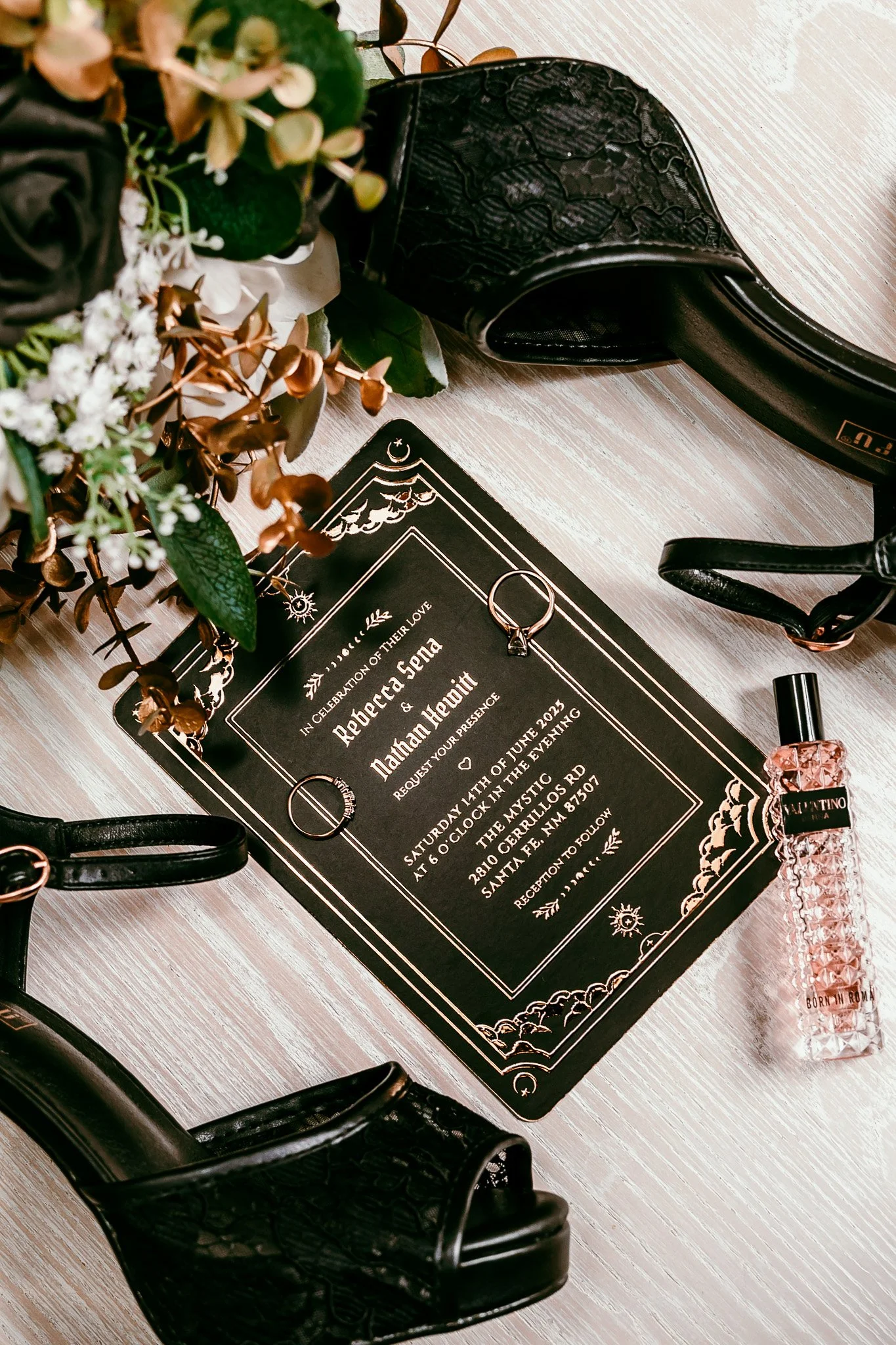 Flat lay of black wedding invitation, bridal heels, wedding rings, perfume, and bouquet details for an intimate wedding at The Mystic in Santa Fe, New Mexico.