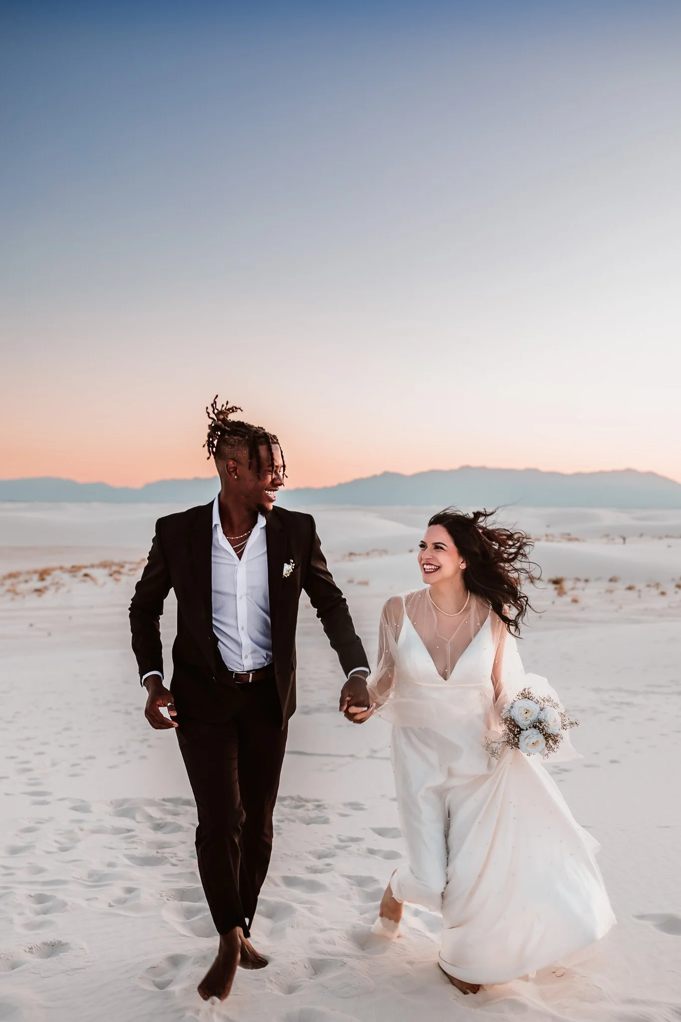 Newly married couple laughing while walking hand in hand through the white dunes at sunset in White Sands National Park, New Mexico.