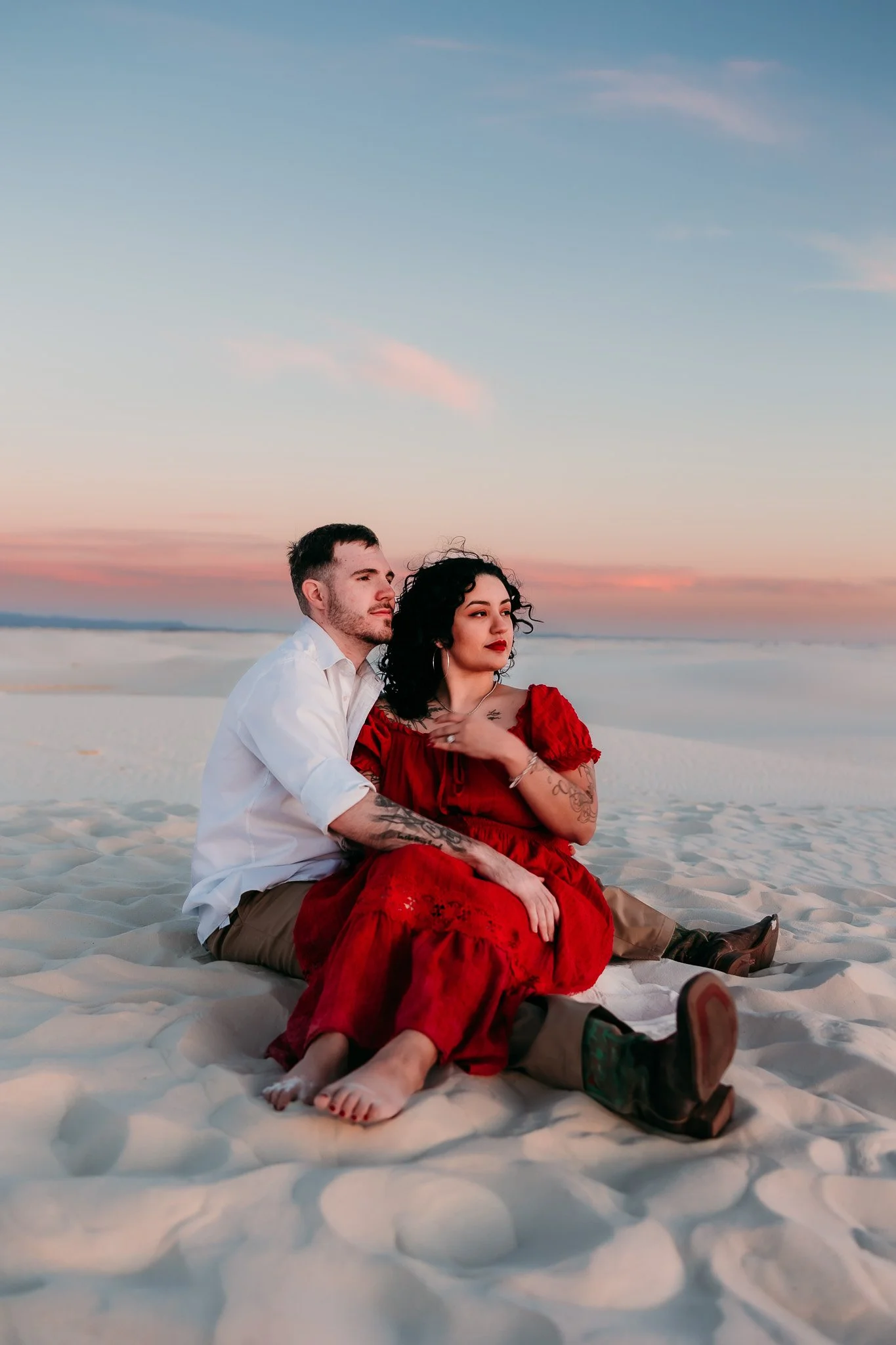 Engaged couple sitting together on the white sand dunes at sunset during a romantic White Sands National Park engagement session.