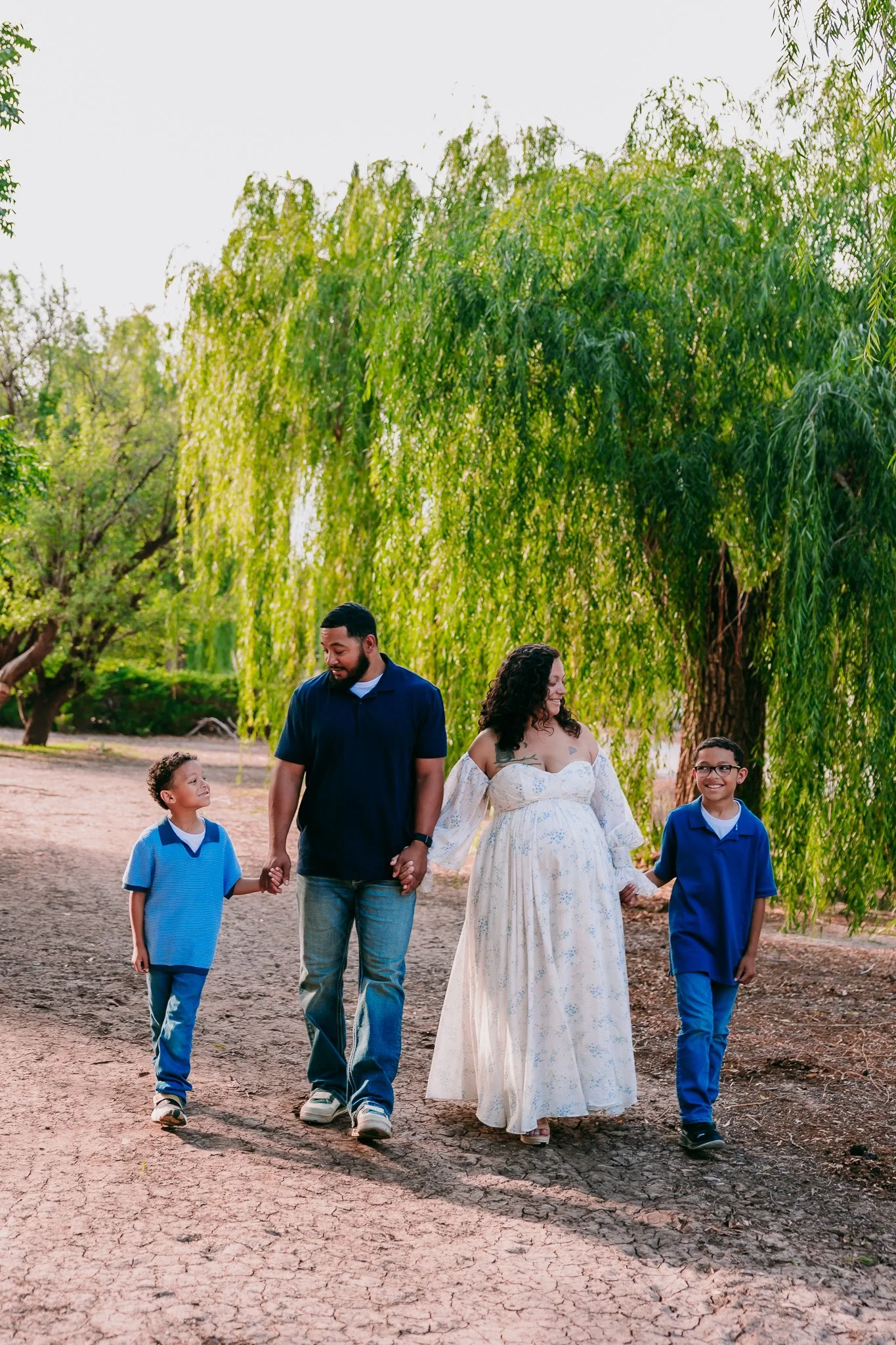 Growing family walking together beneath the willow trees during an outdoor maternity session in Las Cruces, New Mexico.