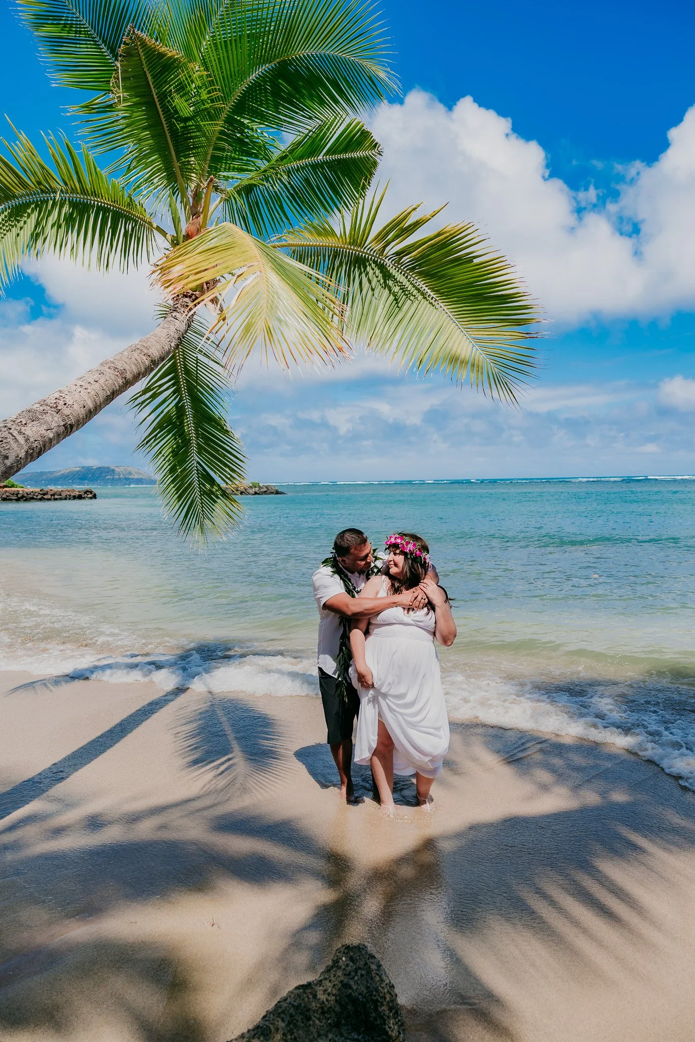 Newlyweds sharing a romantic embrace on the beach with a palm tree overhead during their Hawaii destination wedding.