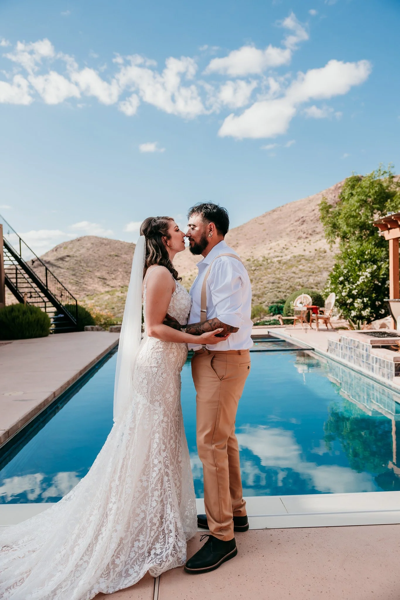 A bride and groom are standing close together, touching noses, by a pool with mountains and a blue sky in the background. The bride is in a lace wedding dress with a long train, and the groom is in a white shirt with suspenders and beige pants.