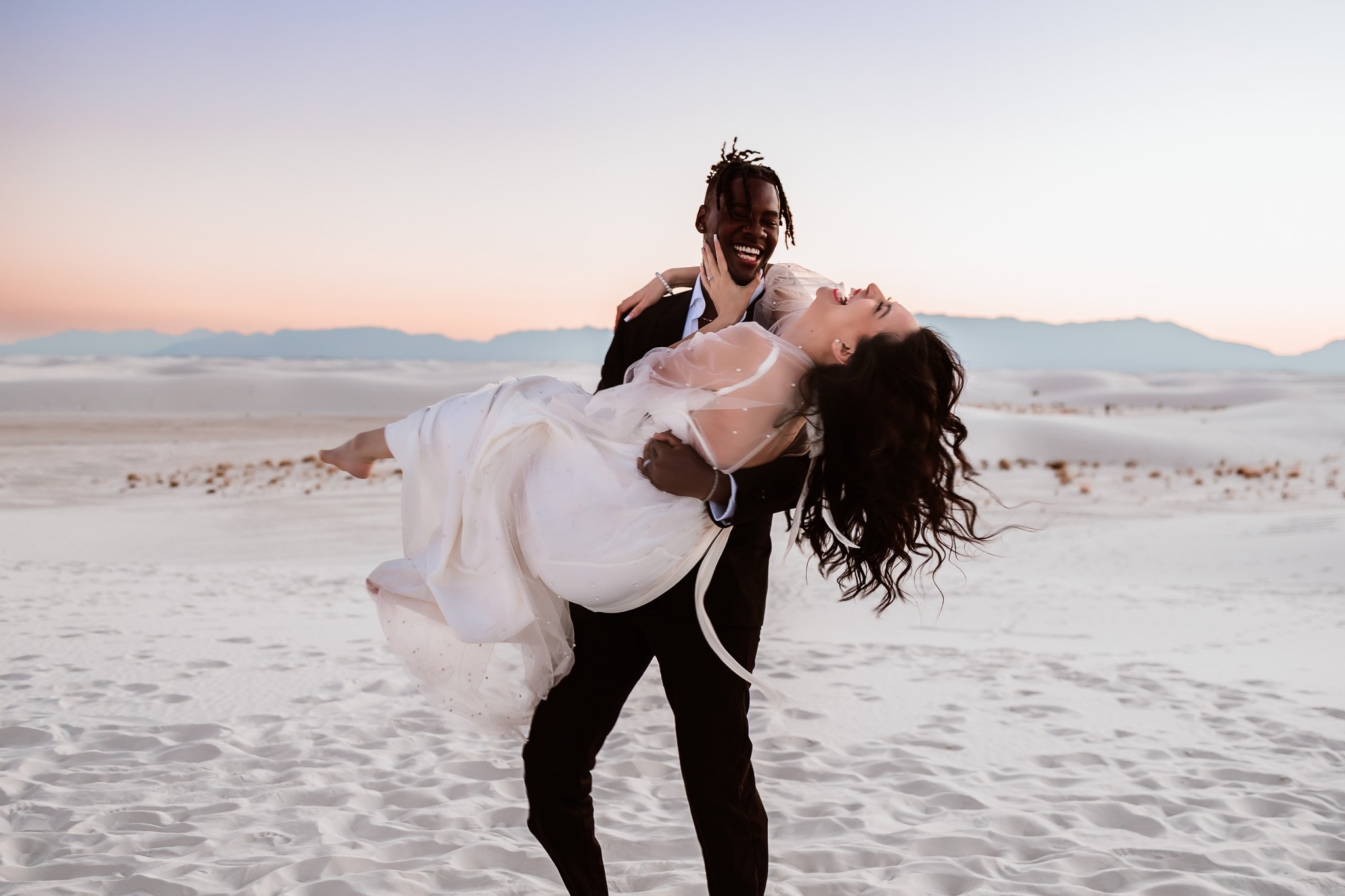 Groom lifting his bride while they laugh together during sunset portraits in the white dunes of White Sands National Park.